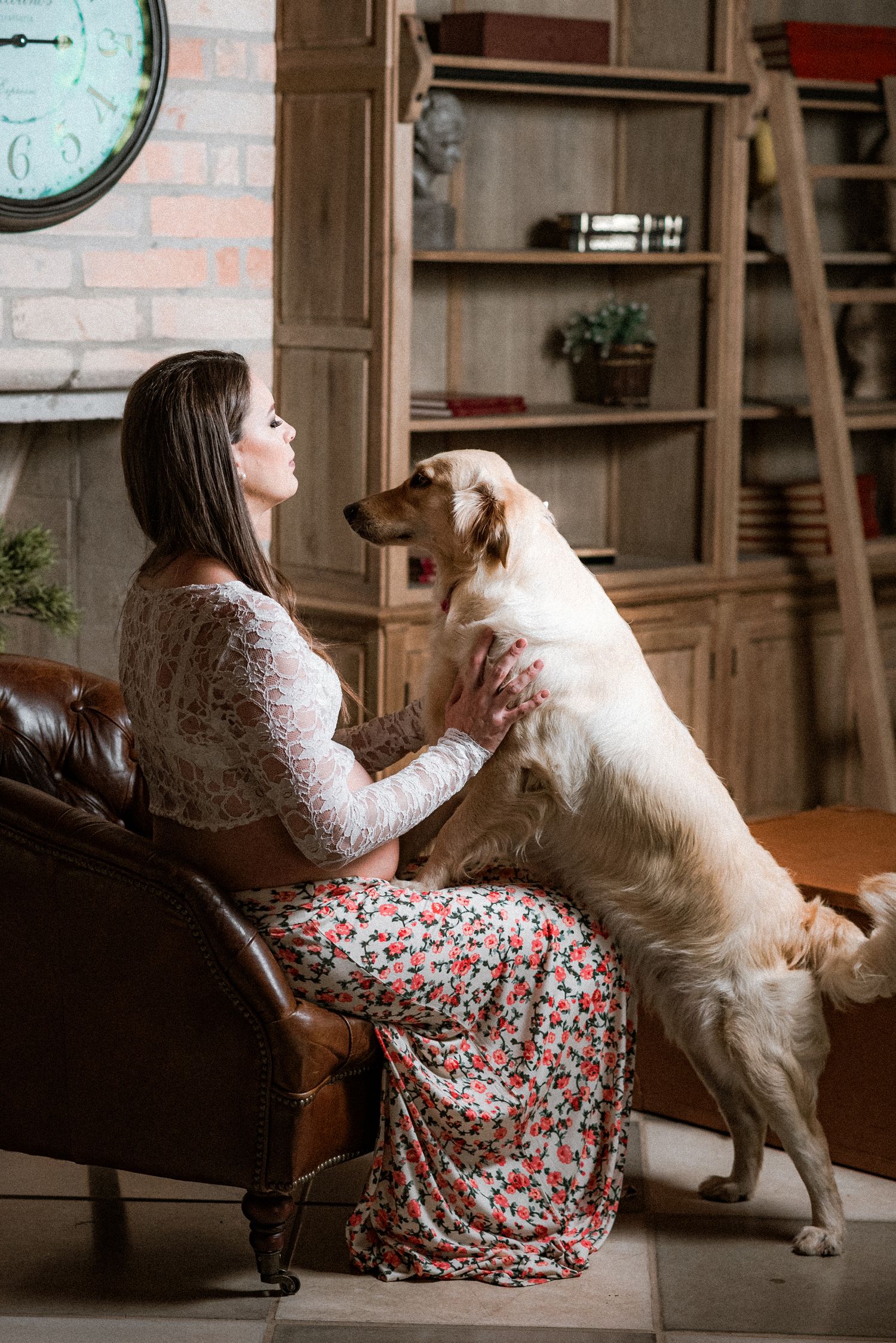 Person in floral dress plays with golden retriever dog on leather chair in rustic room.