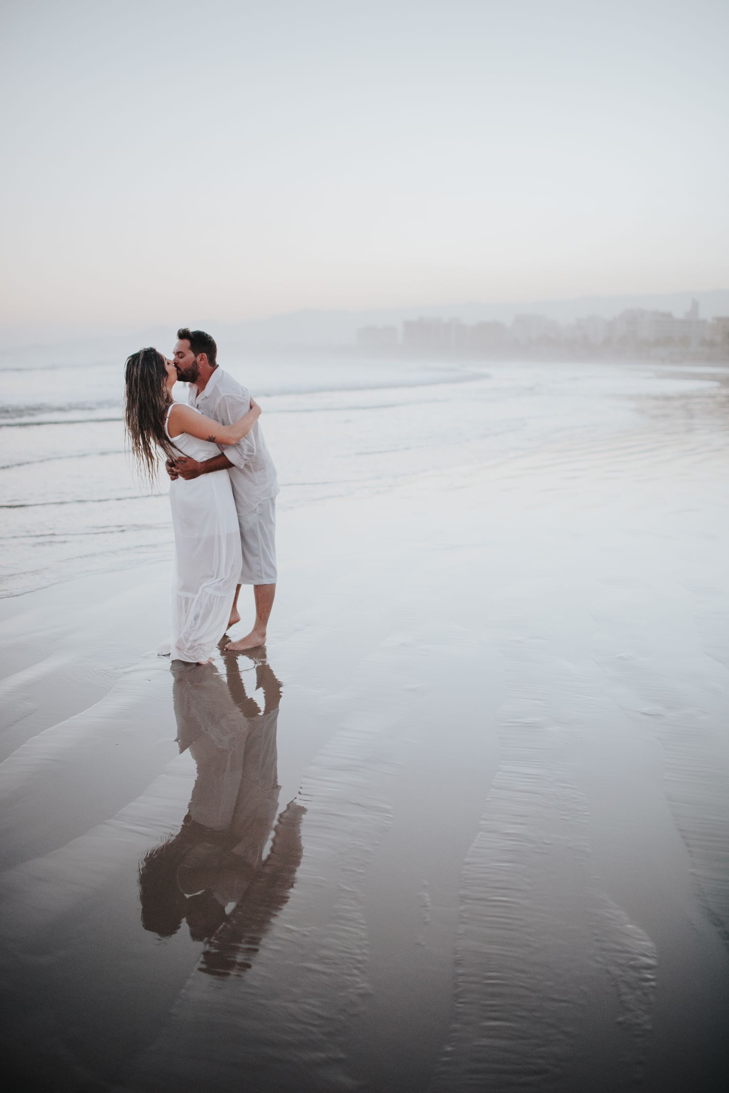 Couple in white clothing dancing together on reflective wet sand at the beach.