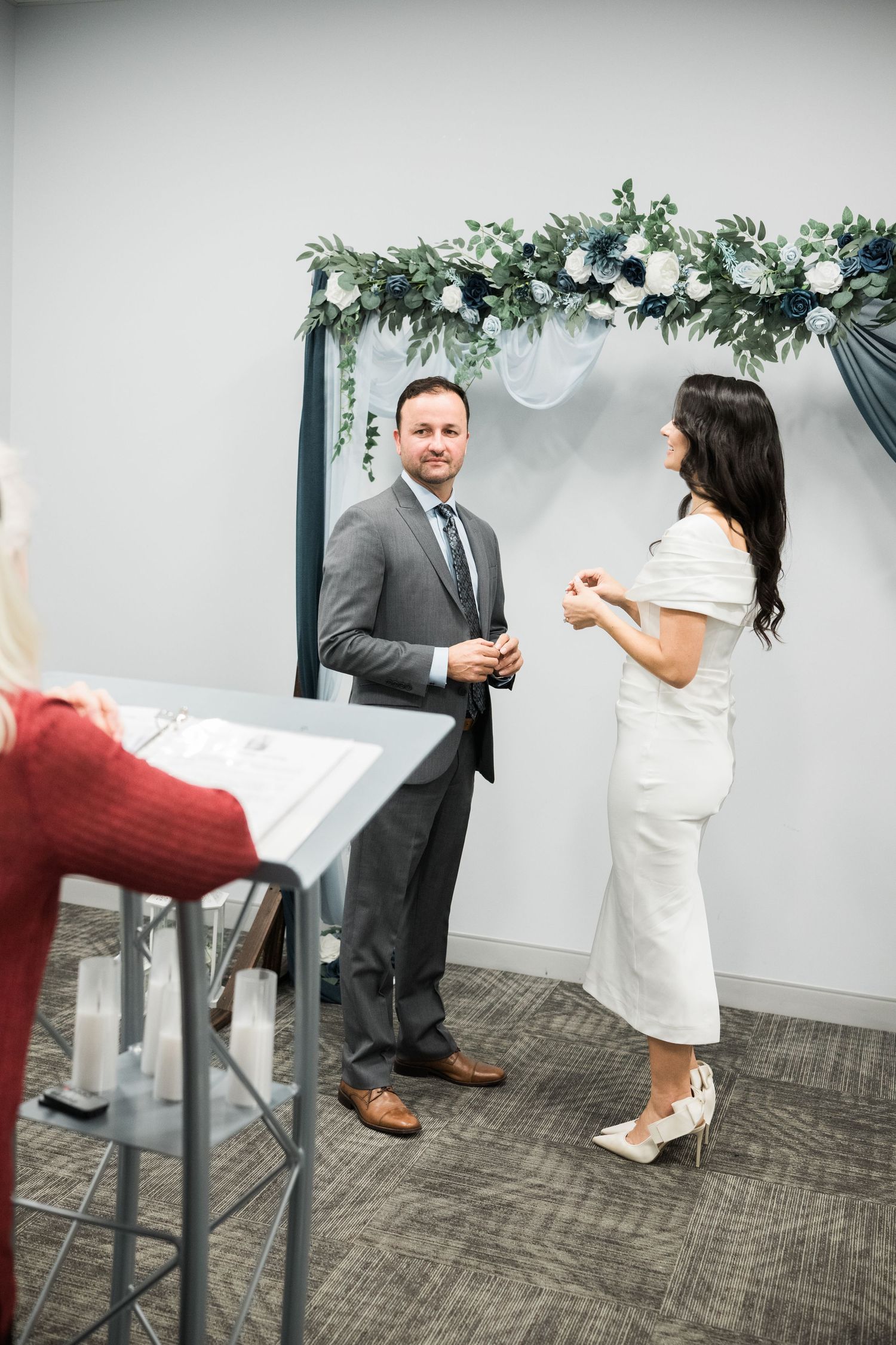 A couple stands together during a wedding ceremony in an office space with floral decor.