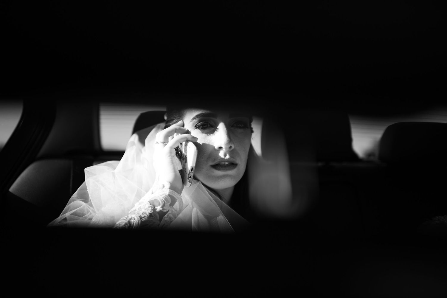Artistic black and white photo through car window showing bride in wedding attire.