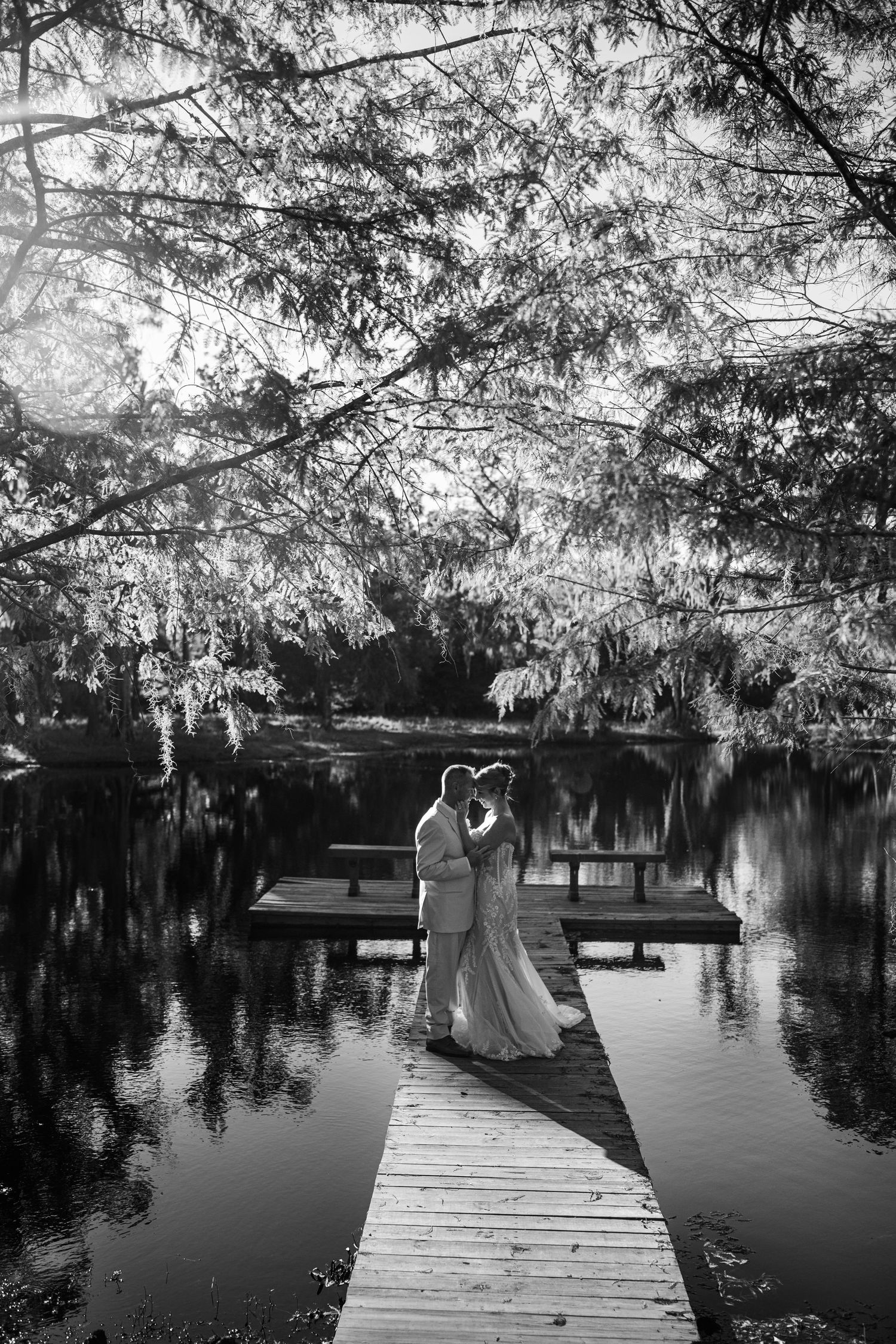 Black and white photo of a wedding couple on a lakeside dock under hanging moss.
