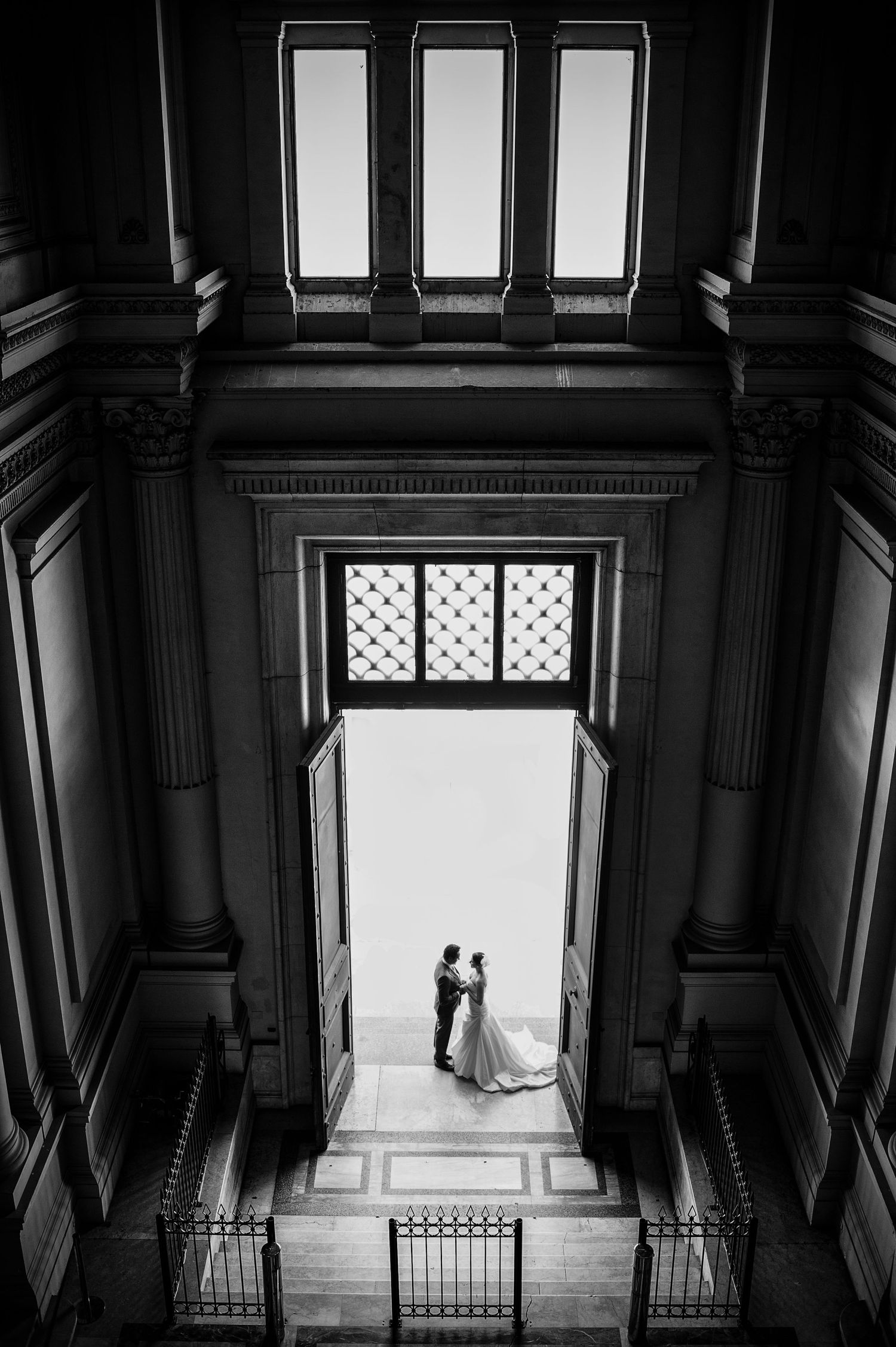 Dramatic black and white architectural photo of a couple in a grand doorway with tall windows above.