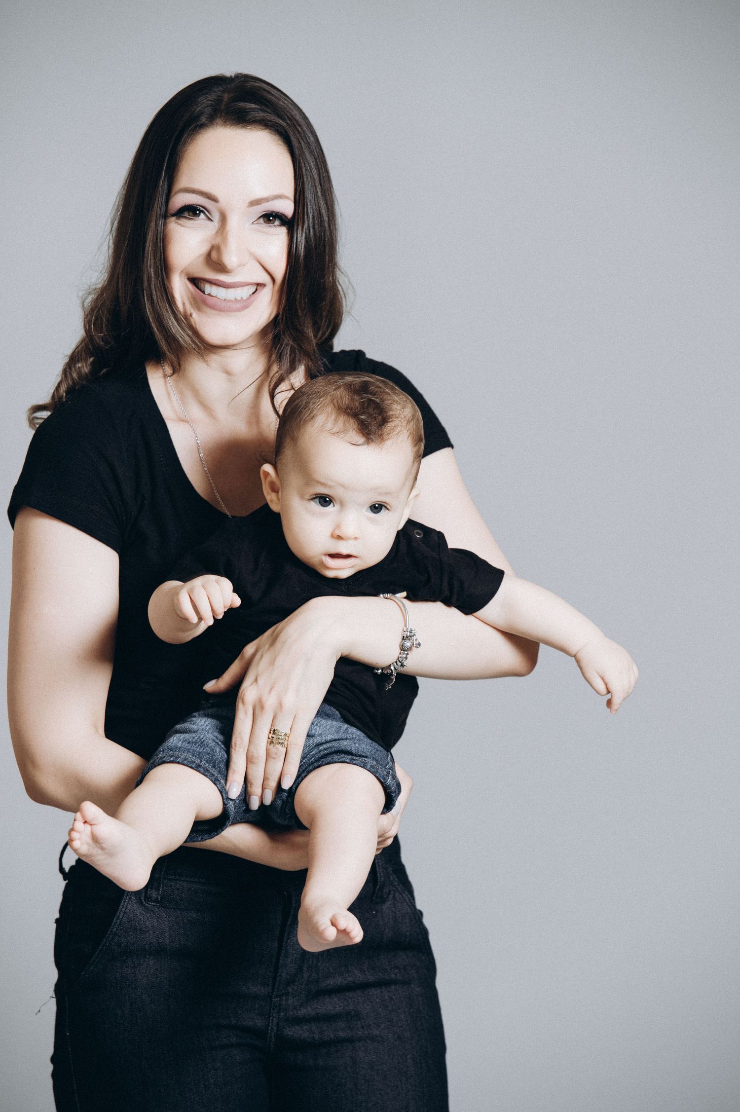 A parent and baby in black clothing pose together against a light gray studio background.