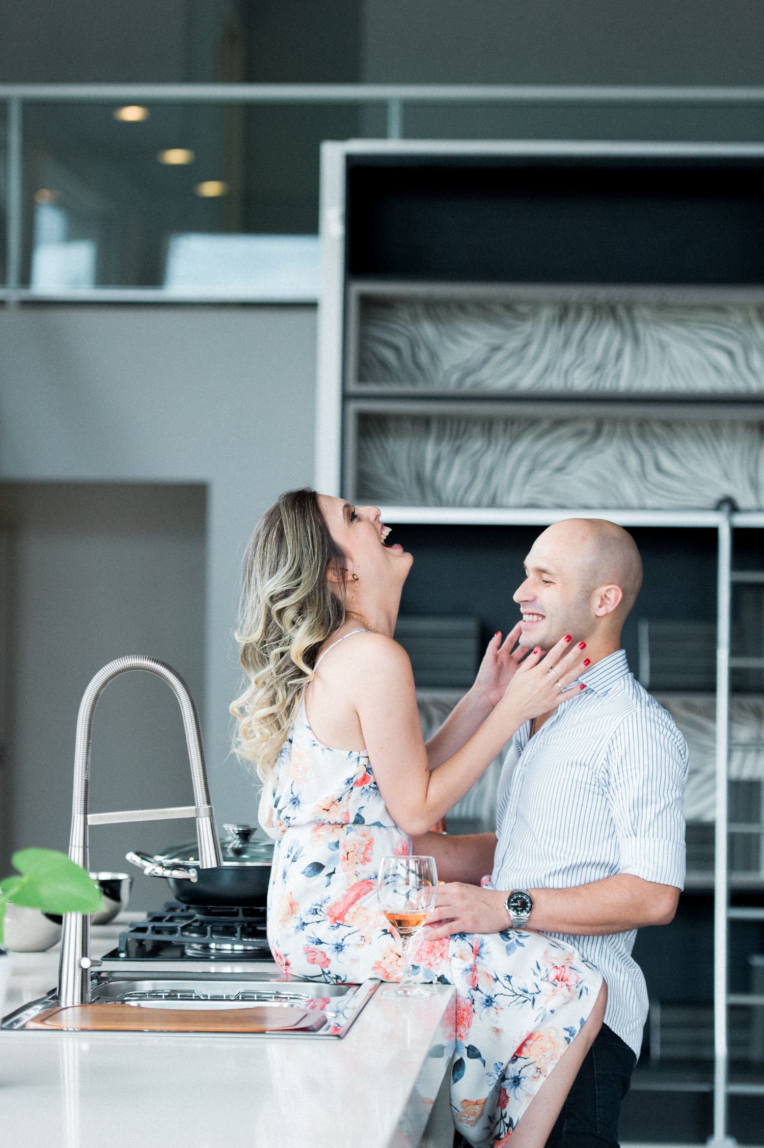 Couple sharing an intimate moment in a modern kitchen with the woman sitting on the counter wearing a floral dress.