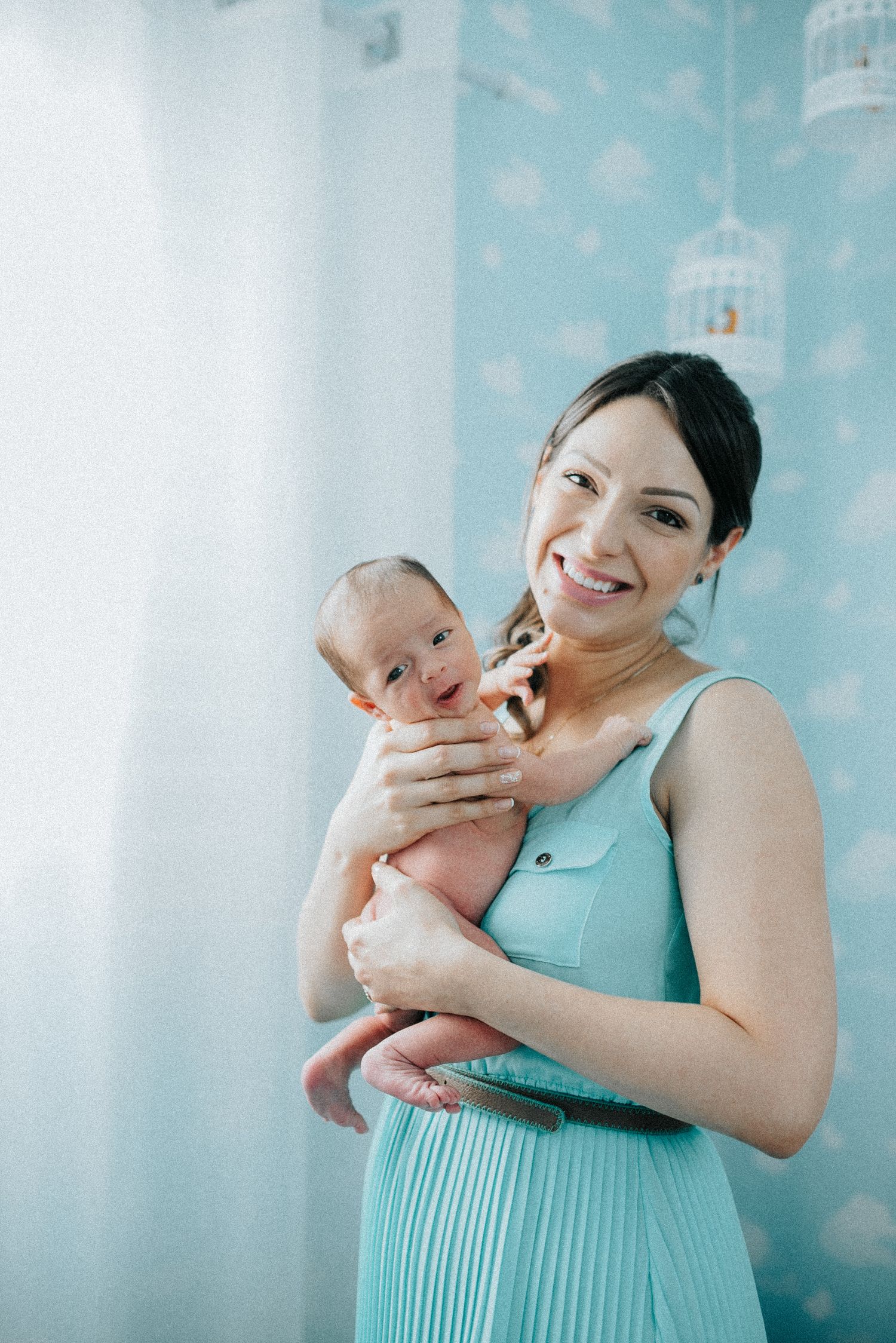 Mother in mint green dress cuddling newborn baby in bright nursery setting.