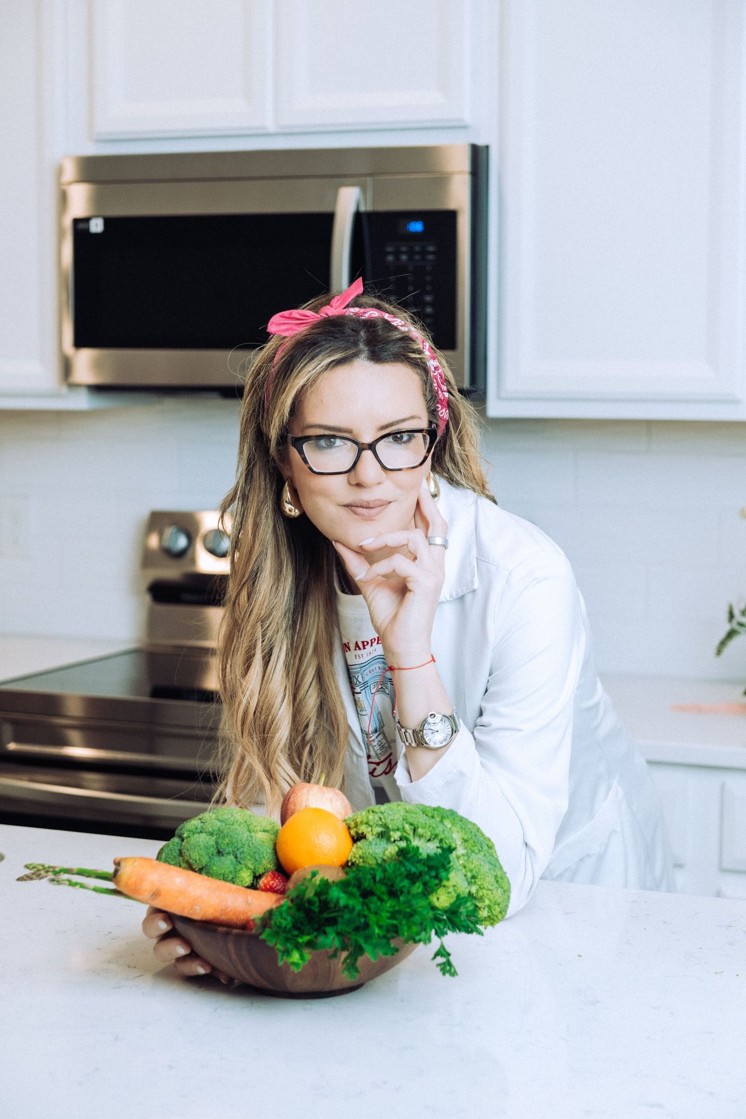 A person in a white coat leans on a kitchen counter with fresh vegetables and produce.