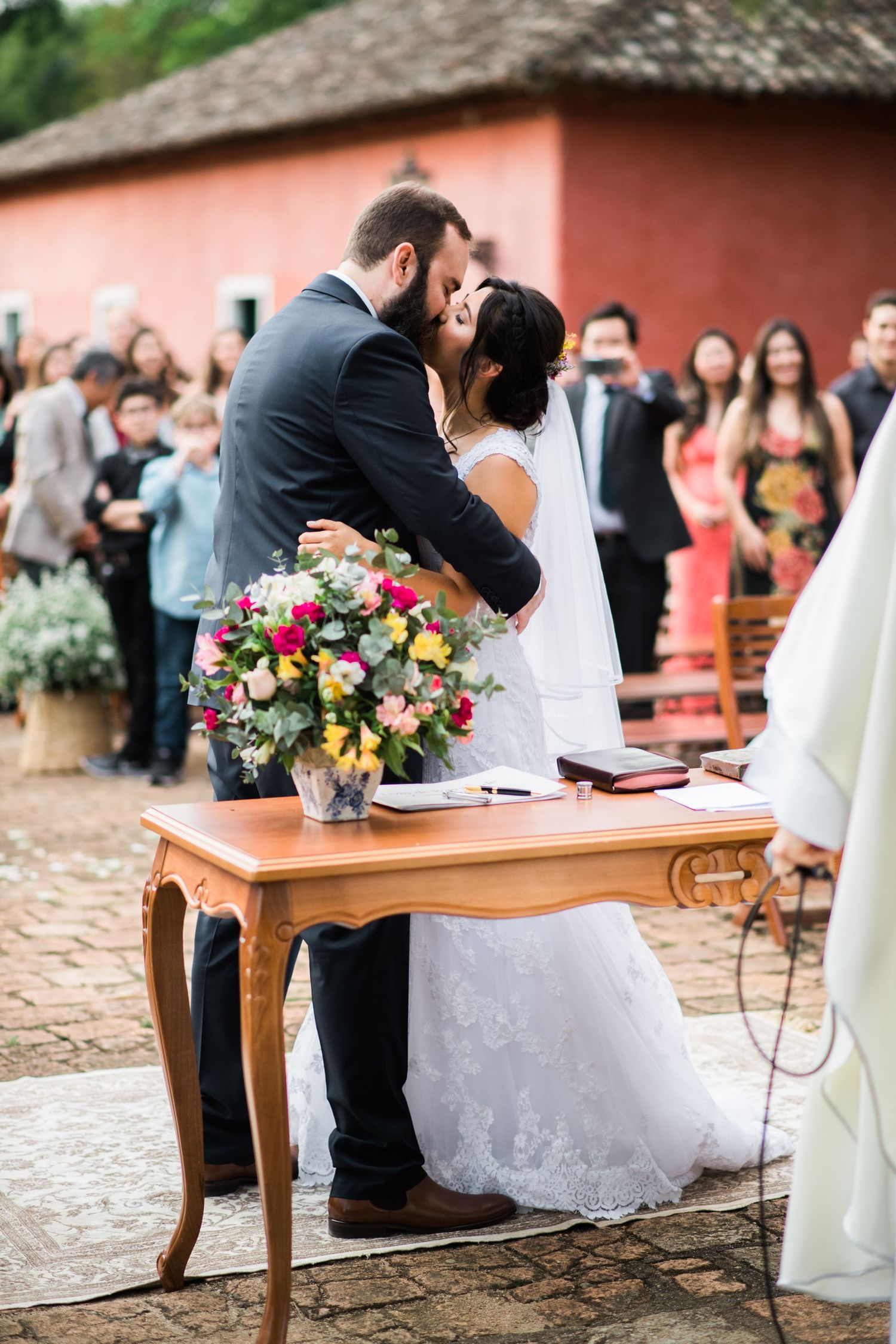A wedding ceremony setup with wooden tables and white chairs against a rustic coral building.