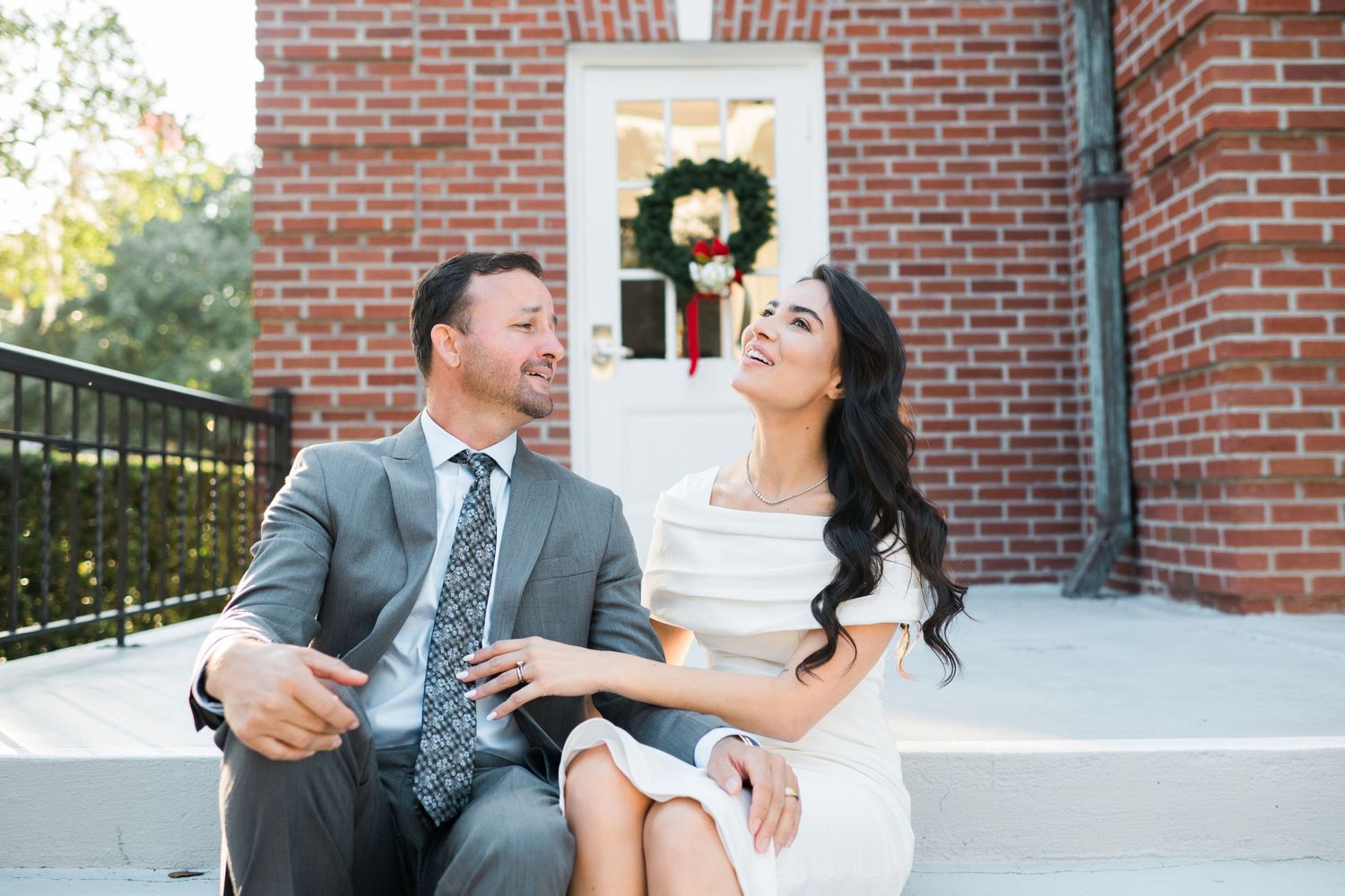 Couple in formal wear sharing a lighthearted moment while sitting on steps of a brick building.