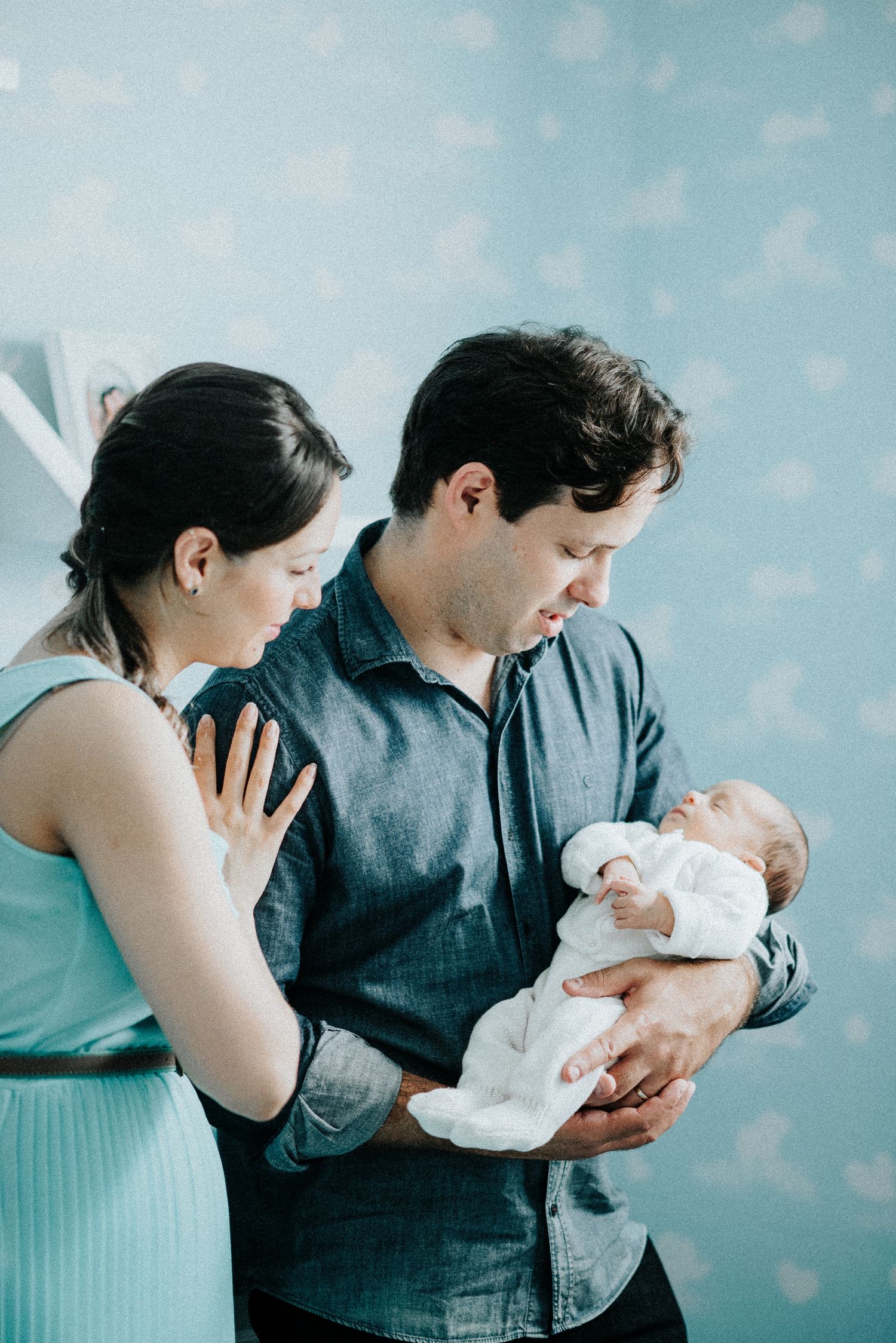 New parents tenderly hold and admire their newborn baby wrapped in white against a blue polka dot background.