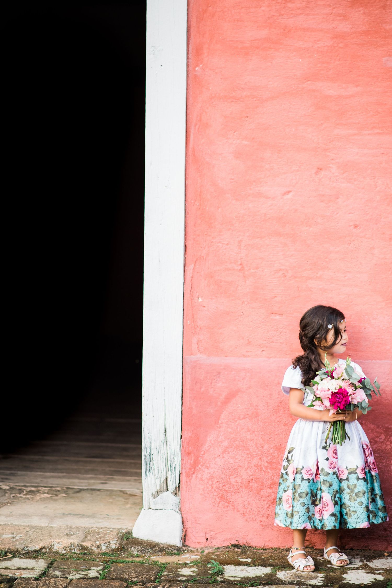 Young flower girl in floral dress poses against pink wall.