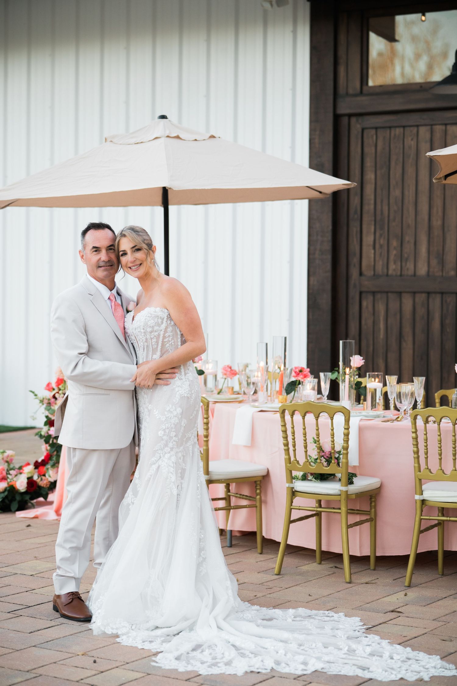 Wedding couple poses by rustic barn wall with elegant table settings and gold chairs in foreground.
