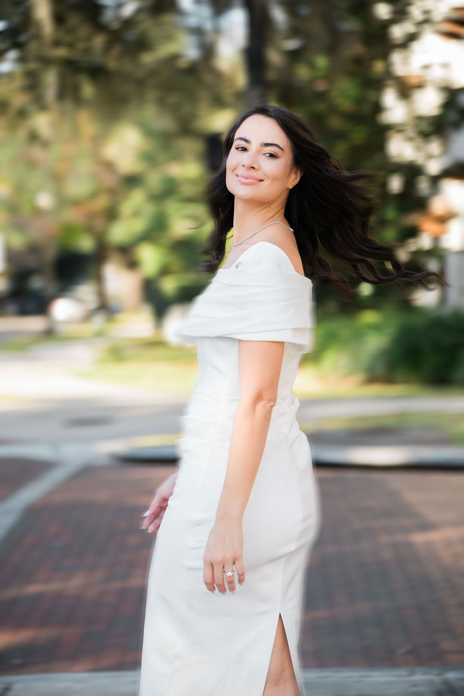 Woman in elegant white dress twirls joyfully outdoors on brick pathway.