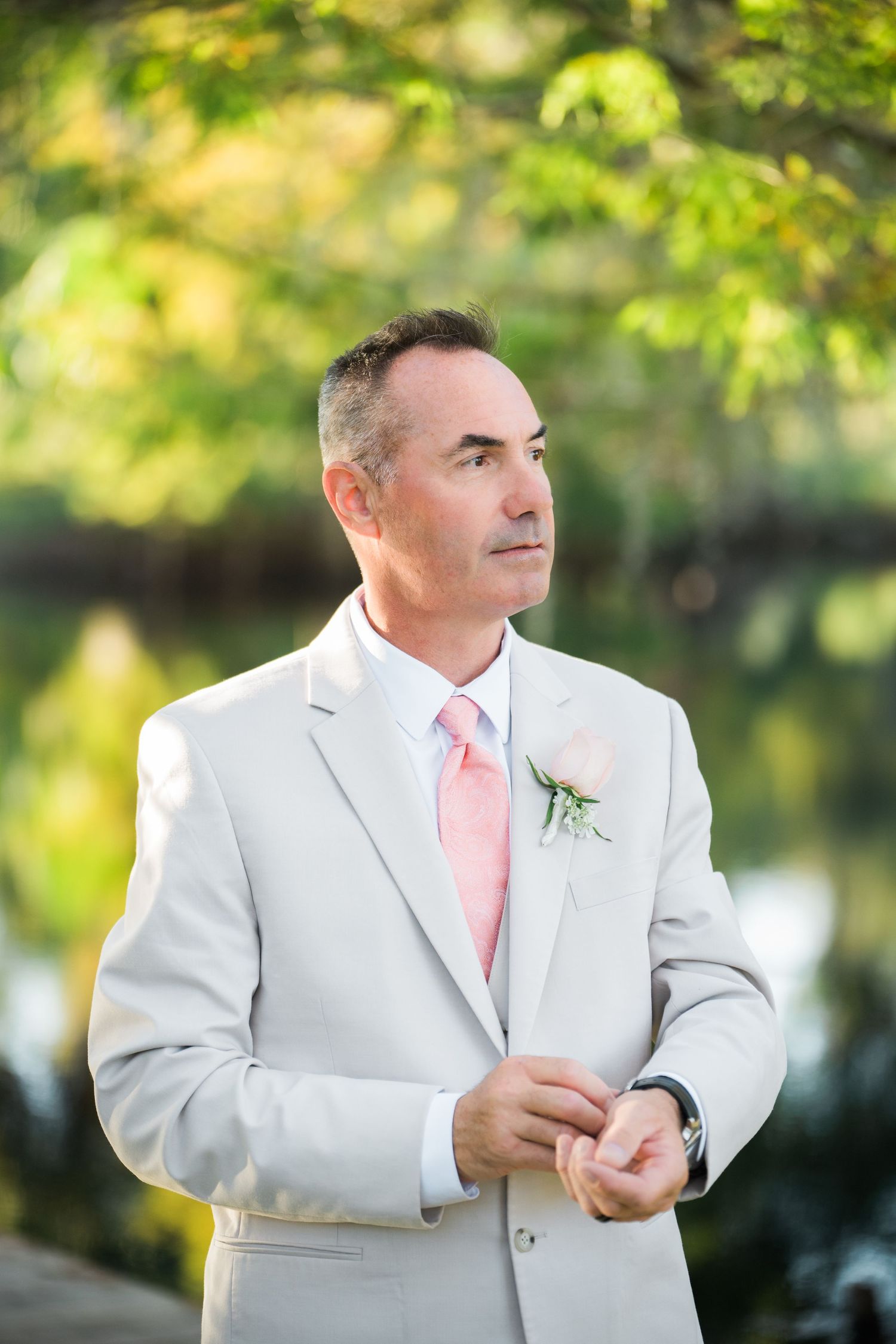 Groom in white suit and pink tie stands against blurred green garden background.