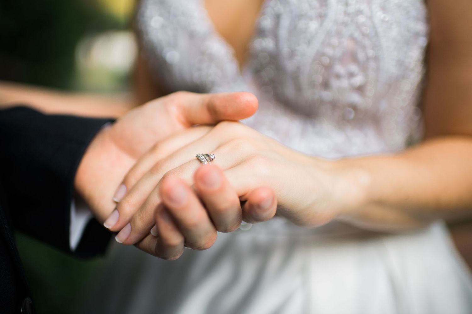 Close-up of newlywed couple's hands with engagement ring and white lace wedding dress in soft focus.