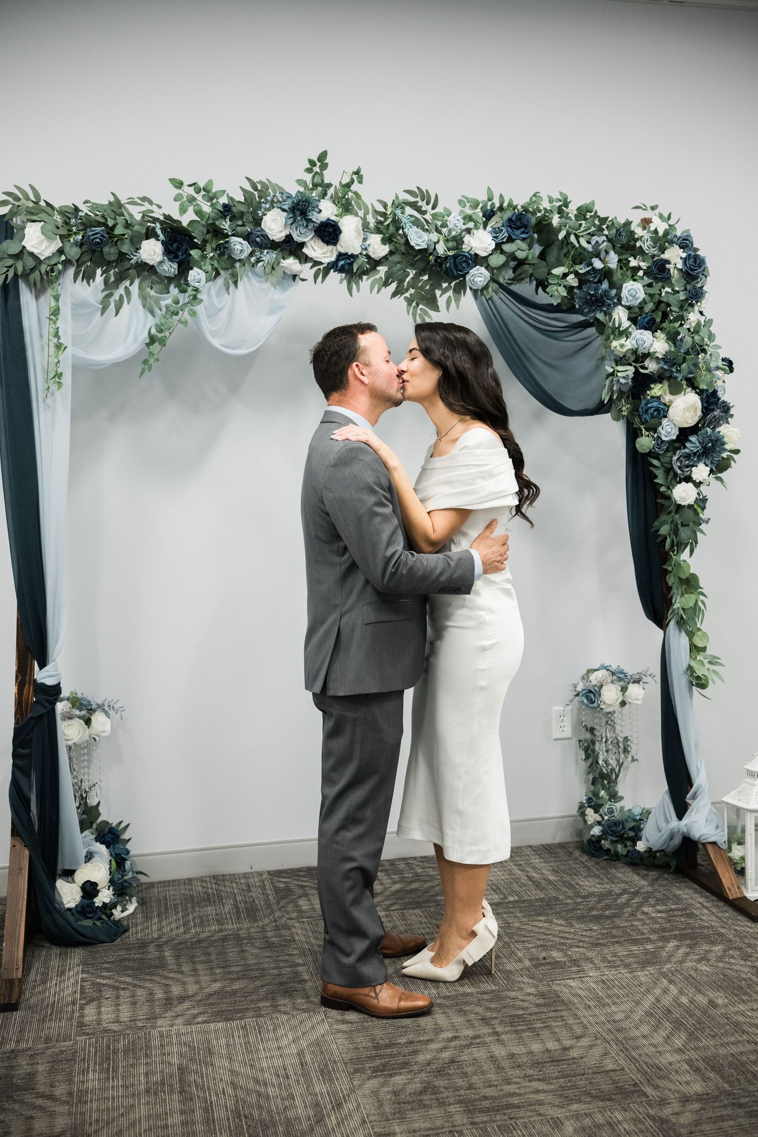 Romantic couple shares intimate moment under floral arch with white roses and greenery.
