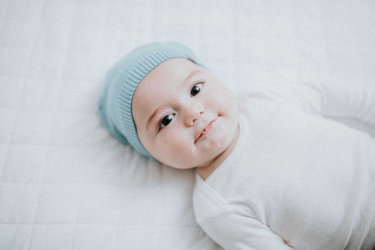 Baby wearing light blue hat lies on white bedding looking at camera.