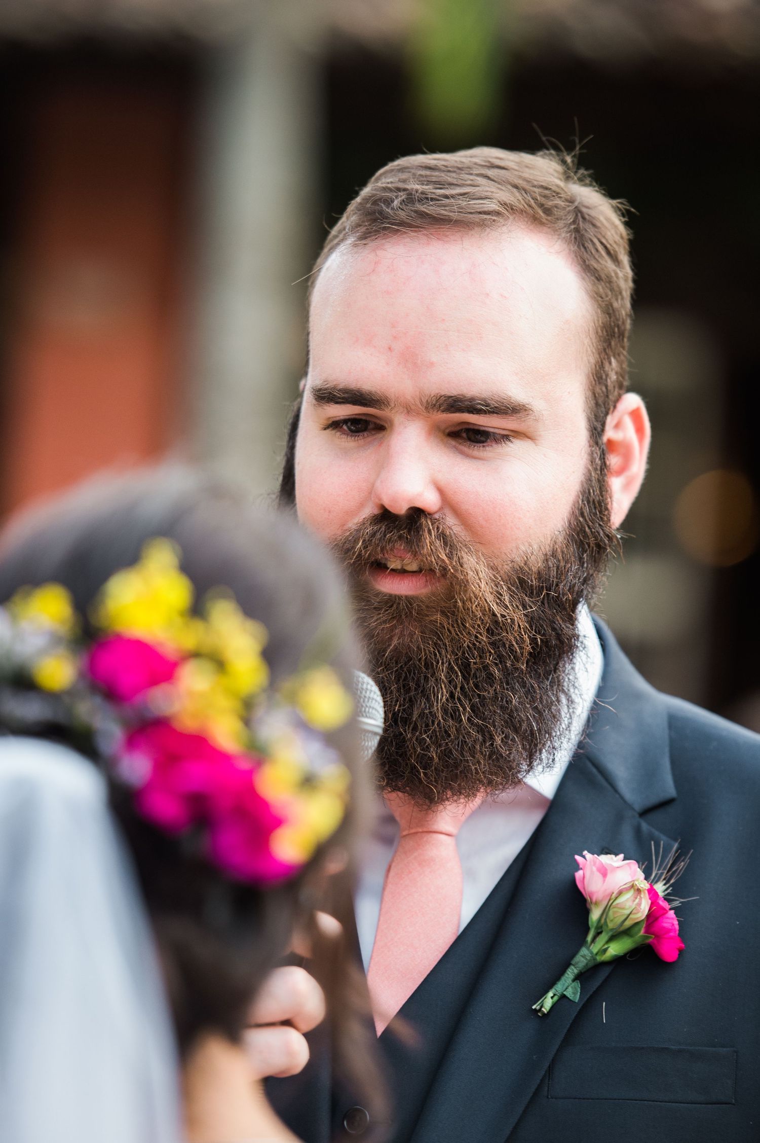 Wedding guest with full beard wearing dark suit and pink floral boutonniere.