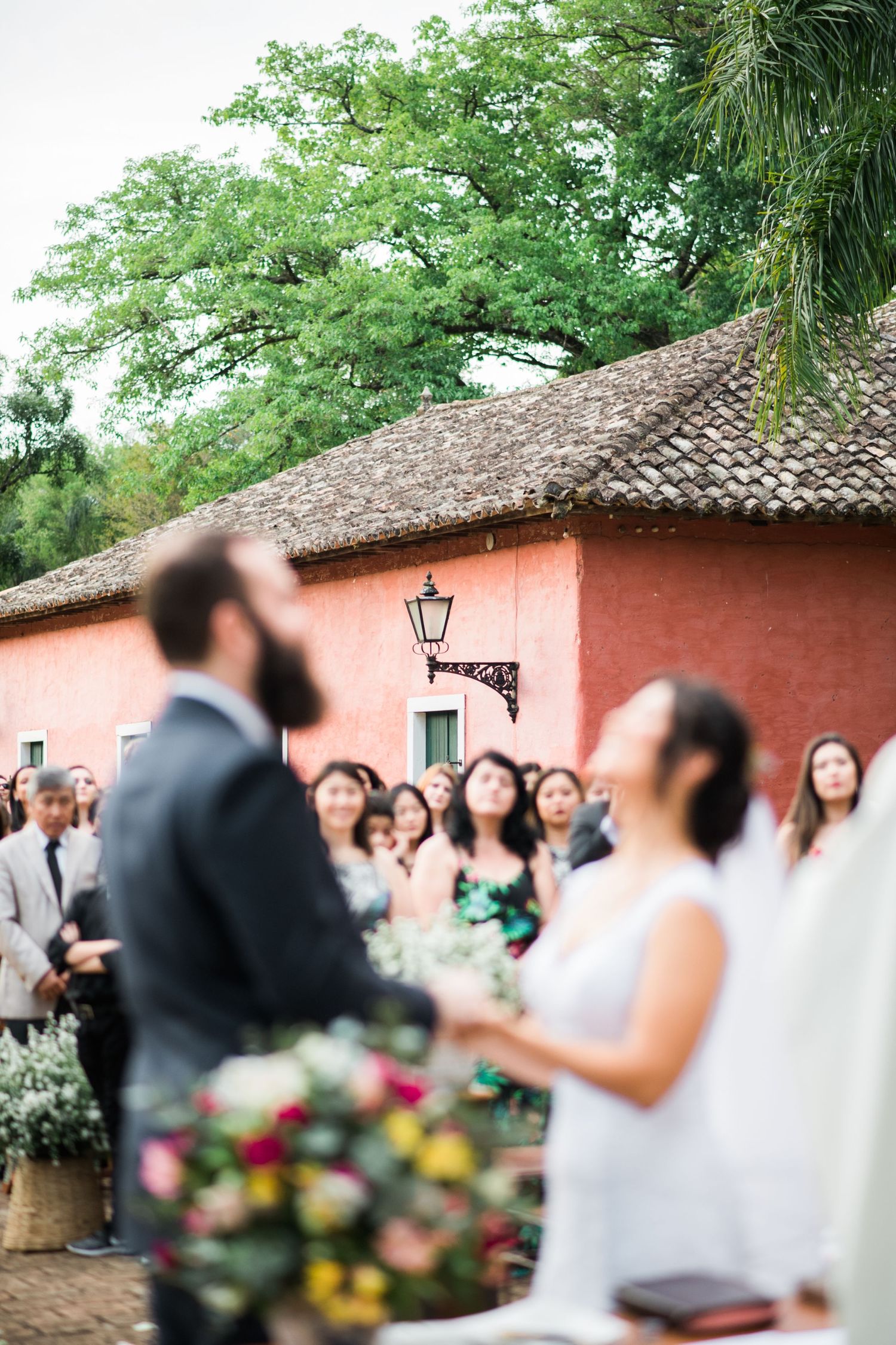 An outdoor wedding ceremony scene with guests gathered around a coral-colored building.
