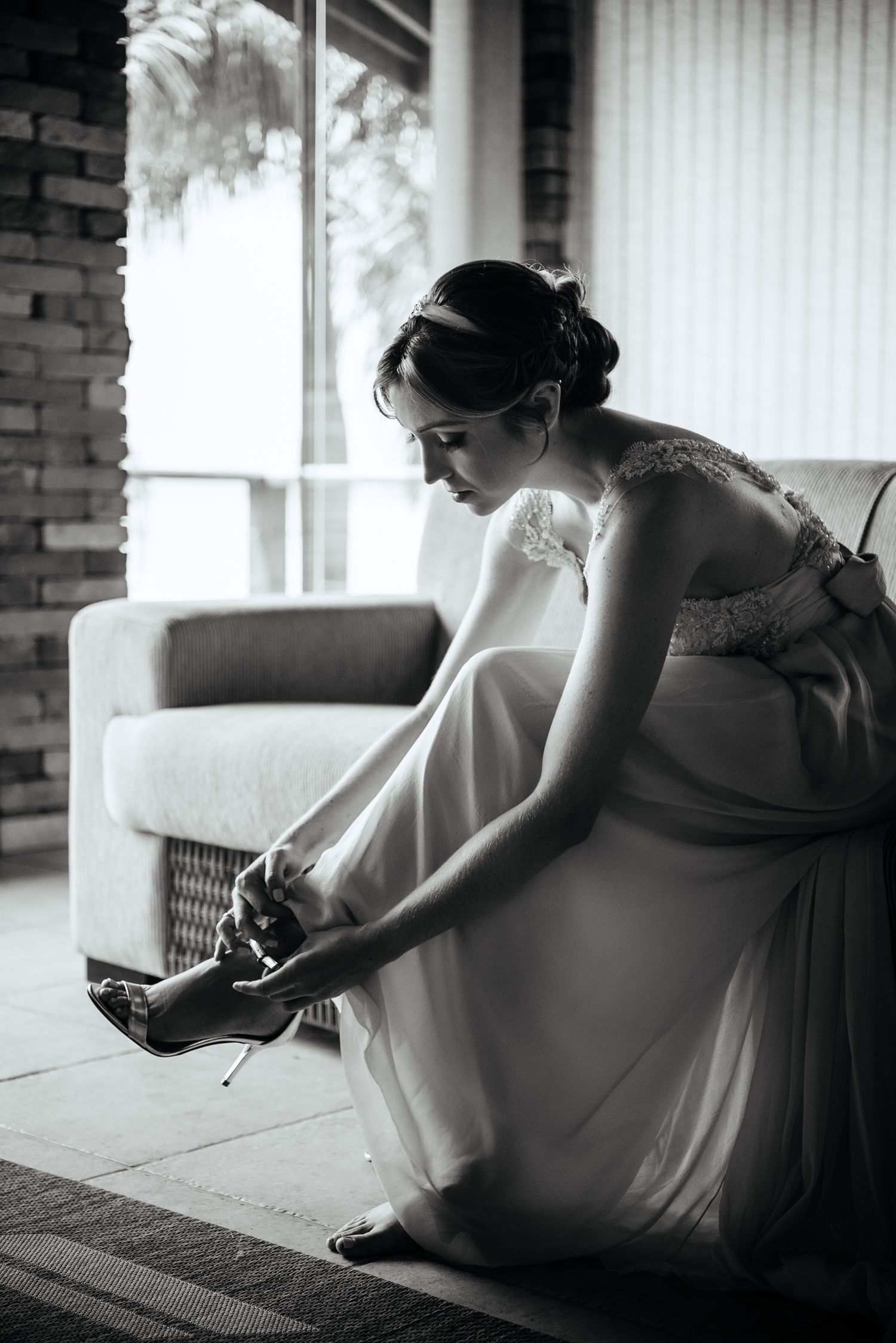 A black and white photograph of a bride putting on her wedding shoes in a sunlit room.