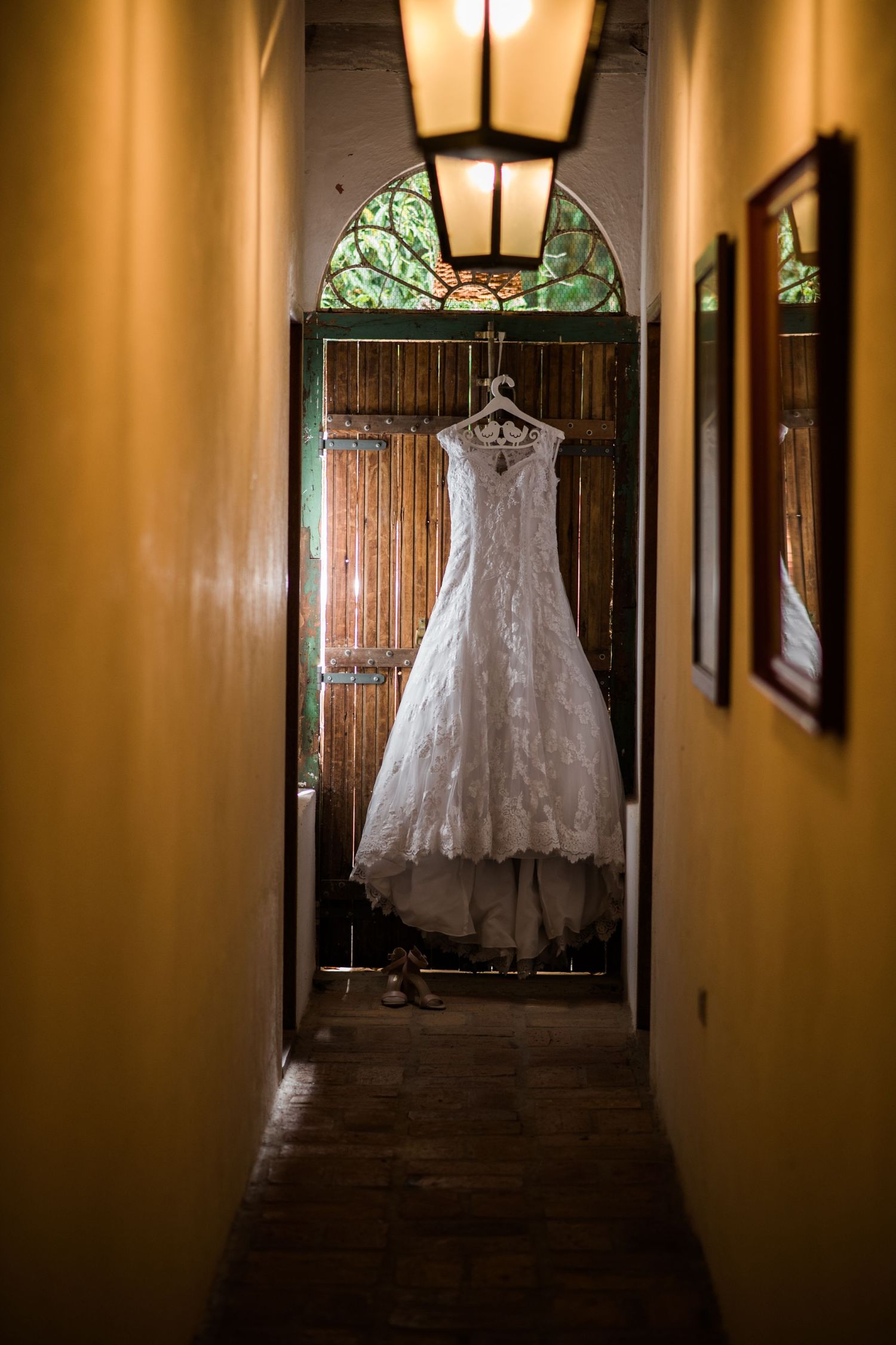 A delicate lace wedding dress hanging in a dimly lit hallway with stained glass and wooden doors.