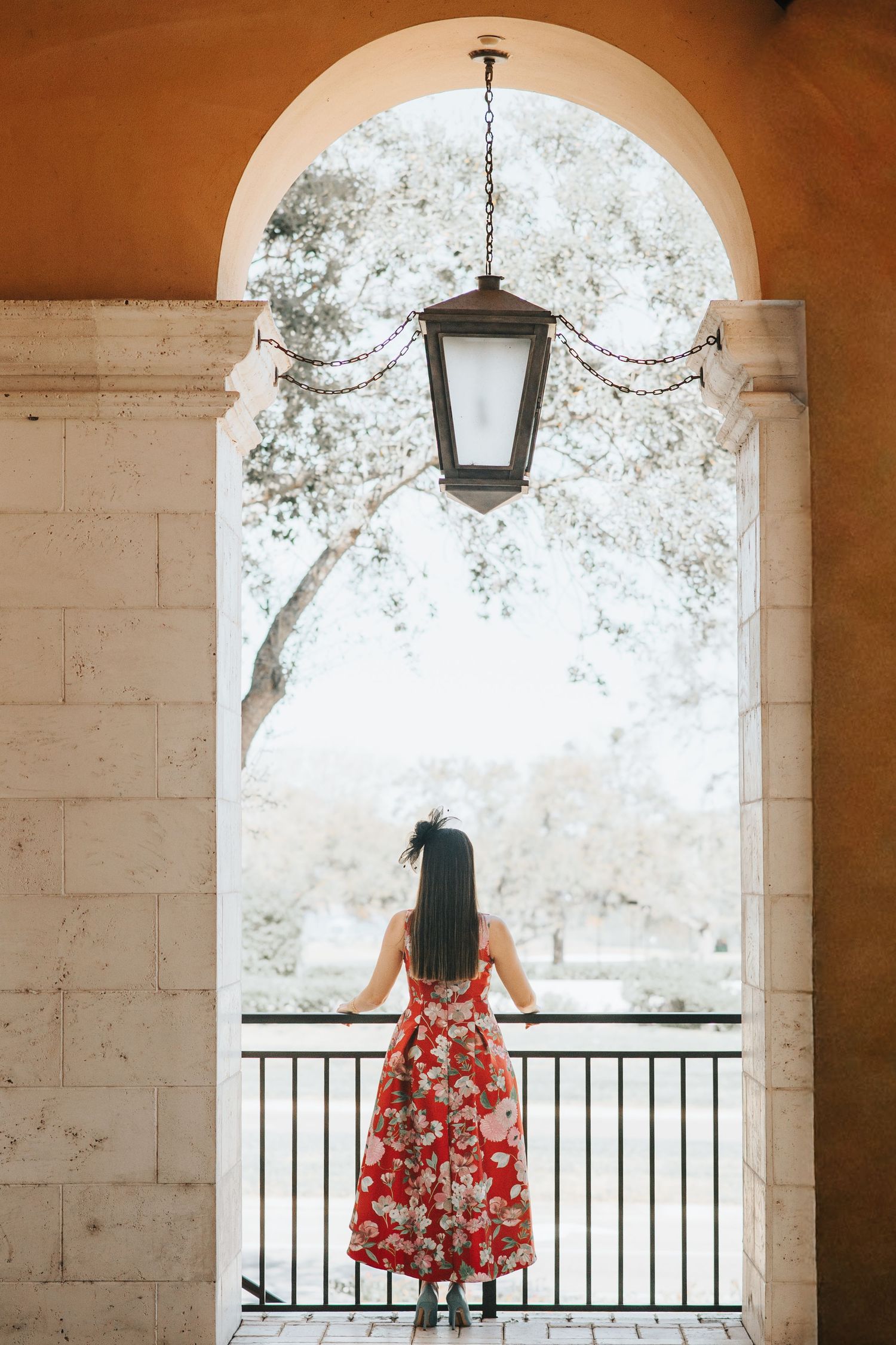 Woman in red floral dress standing under arched doorway looking out at scenic view.
