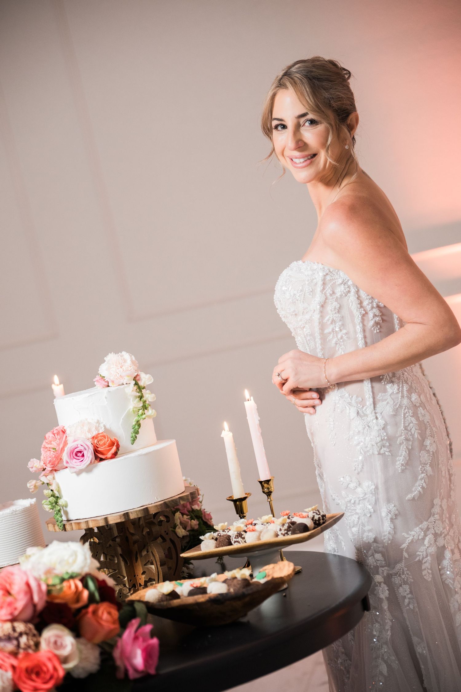 Elegant wedding cake and dessert table display with pink roses and white candles in a light-filled room.