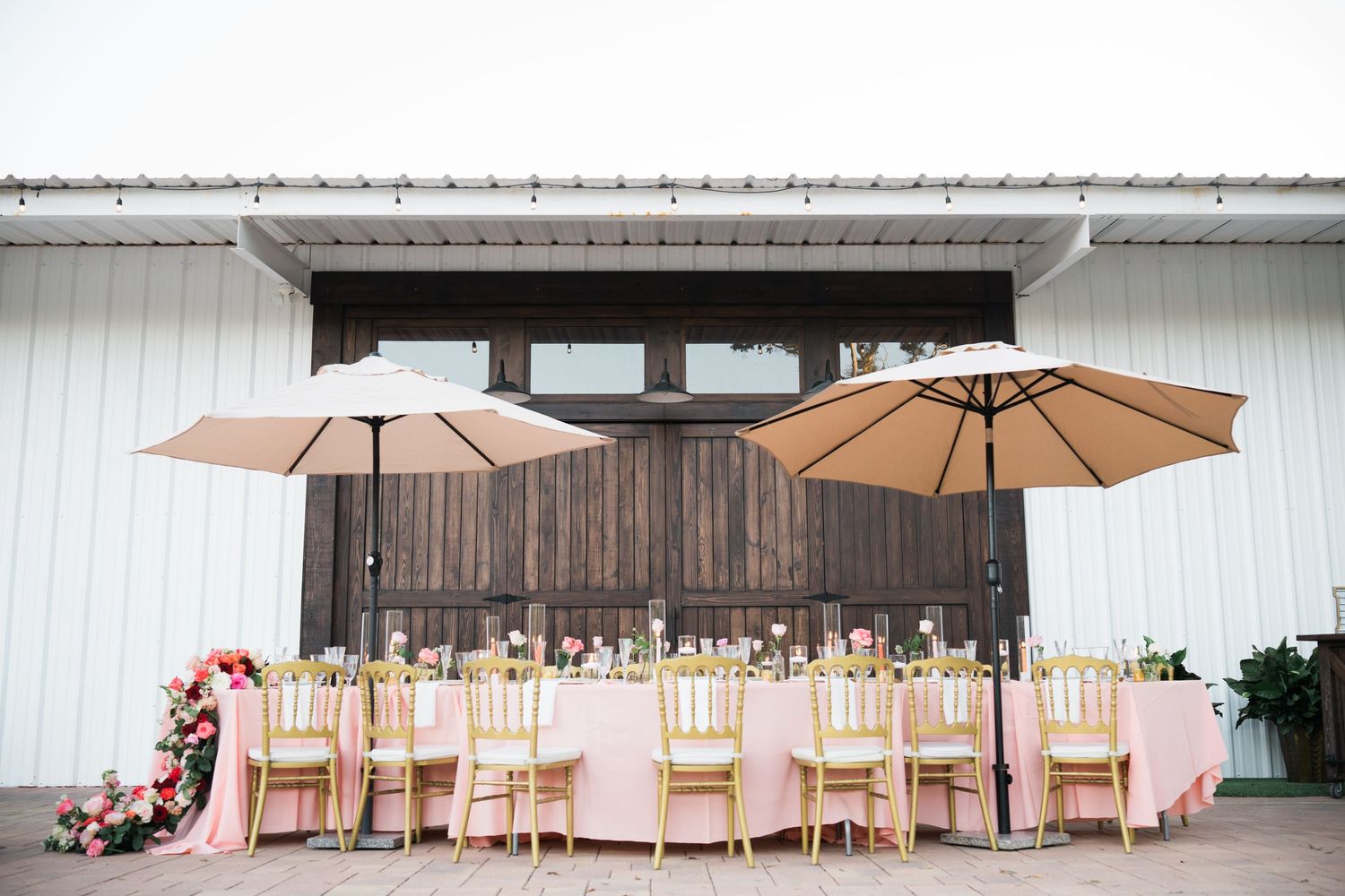 Elegant outdoor wedding reception area with pink linens and beige umbrellas against white barn backdrop.