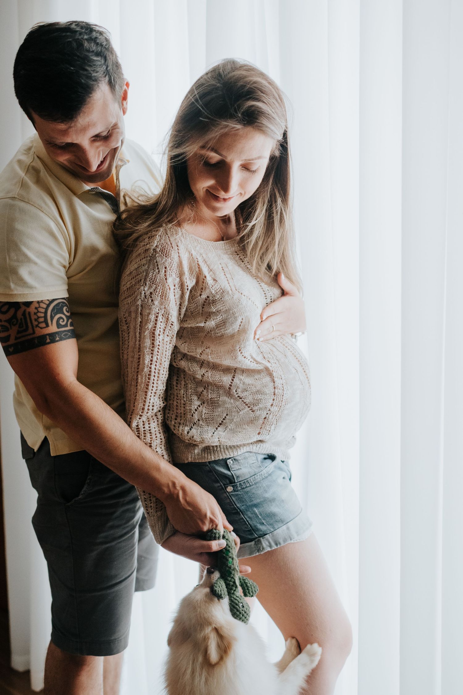 Maternity photos showing couple with small toy props against white curtain backdrop.