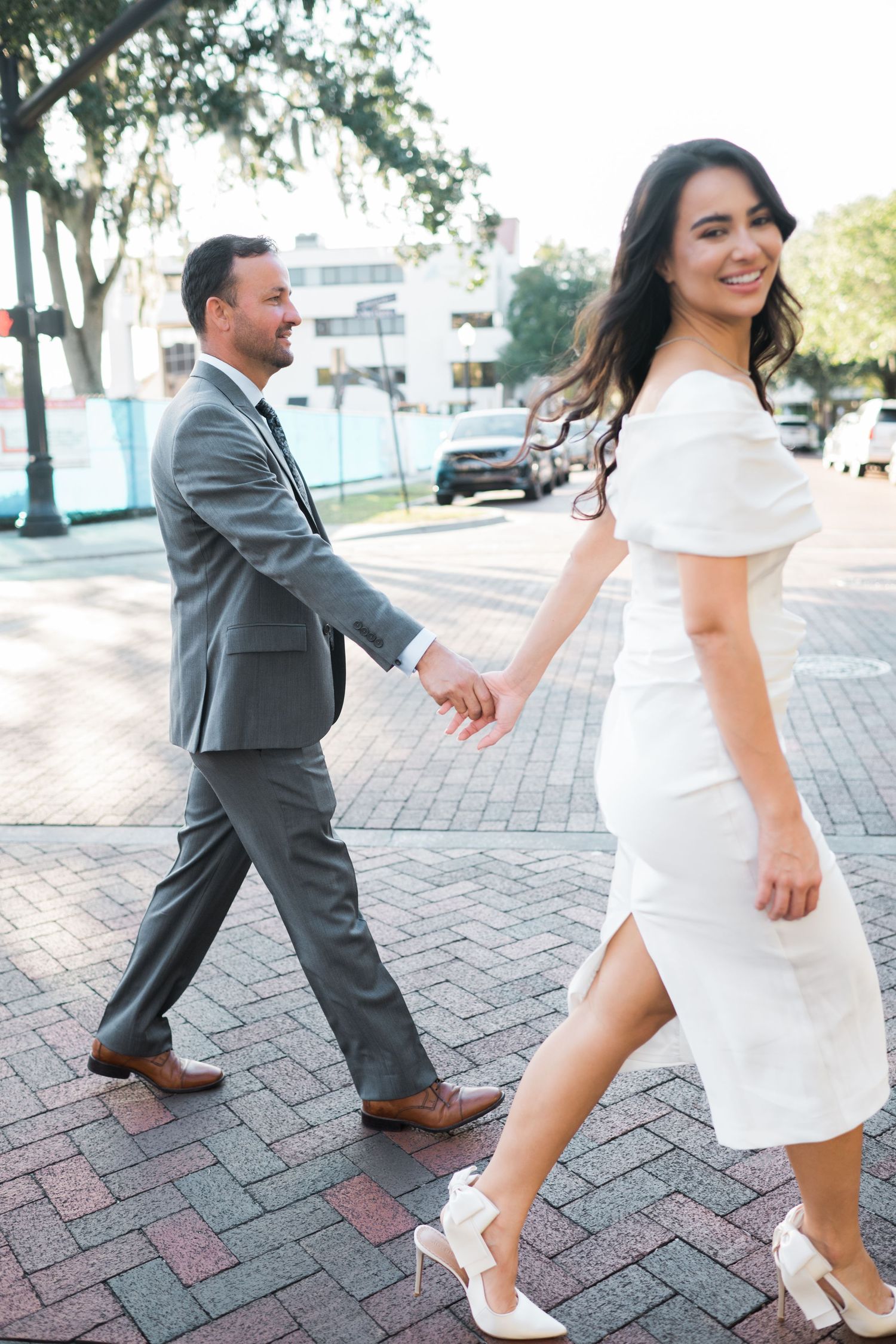 Couple in formal attire walking hand in hand on a brick paved street.