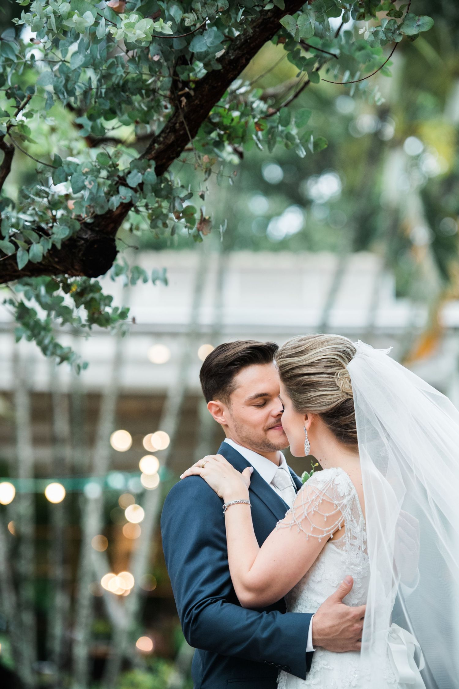 Romantic wedding moment under green trees with string lights in the background.