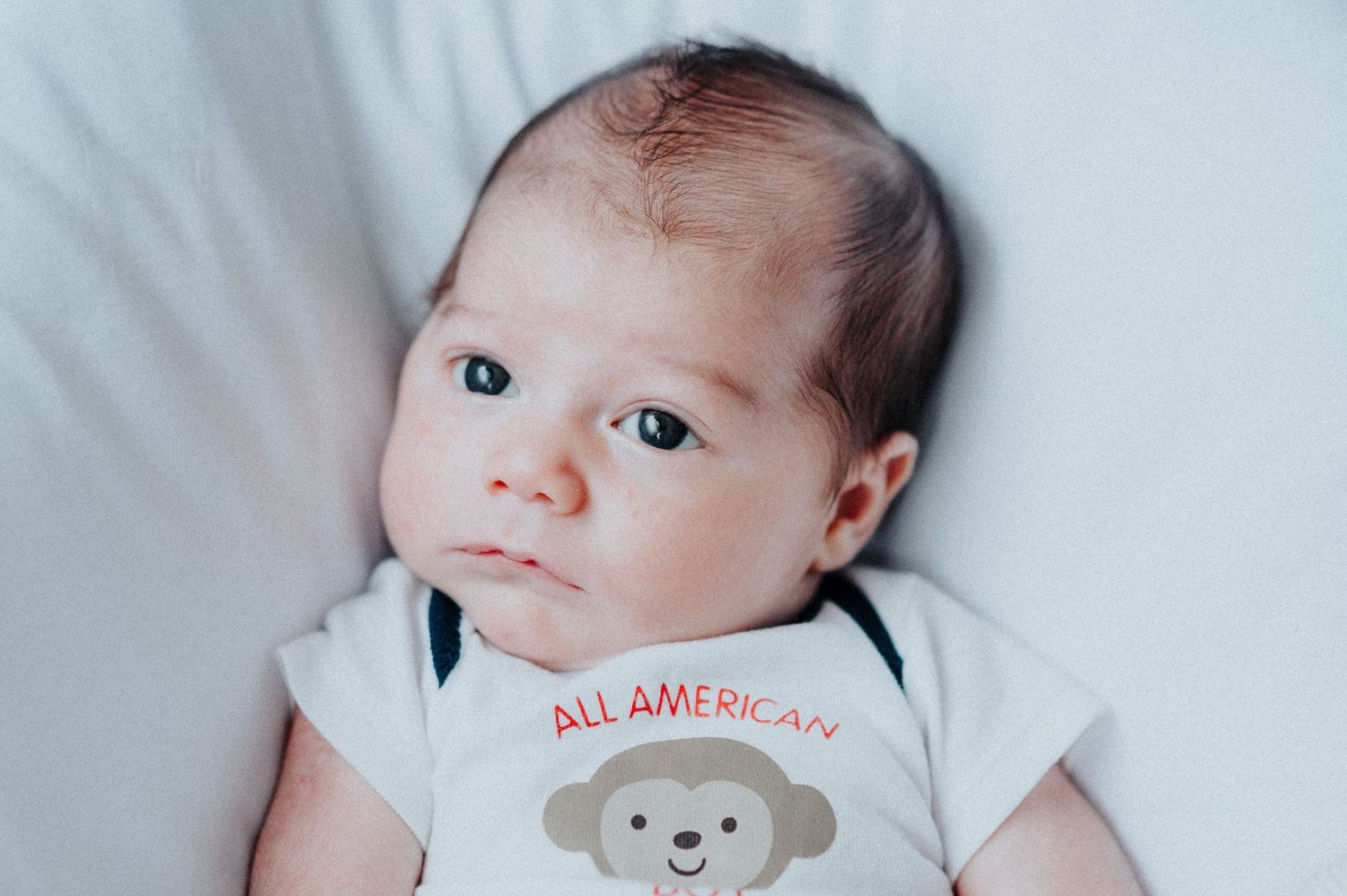Baby wearing white onesie with 'All American' text lying on light background.