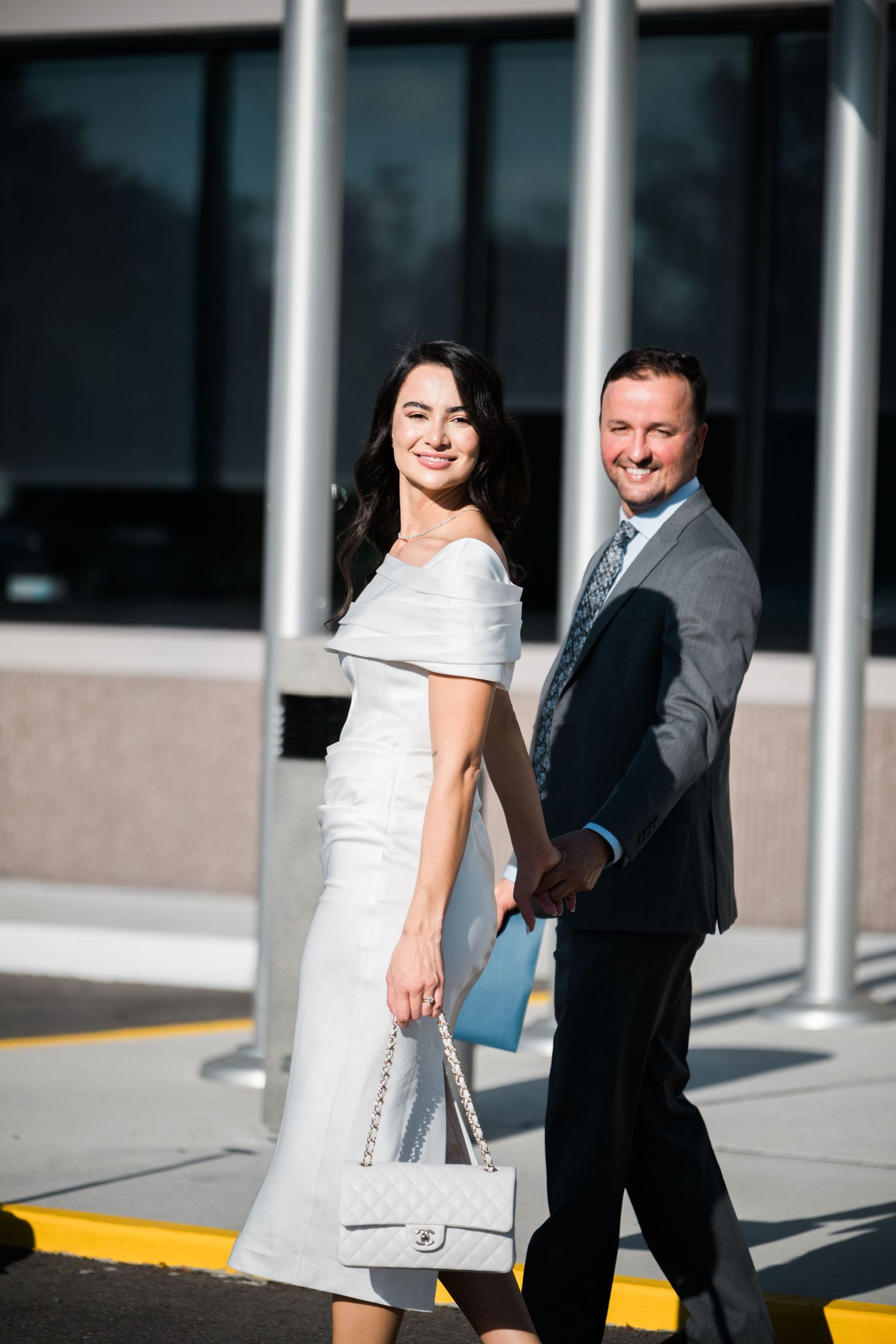 Couple in formal attire poses playfully in front of modern building with columns.