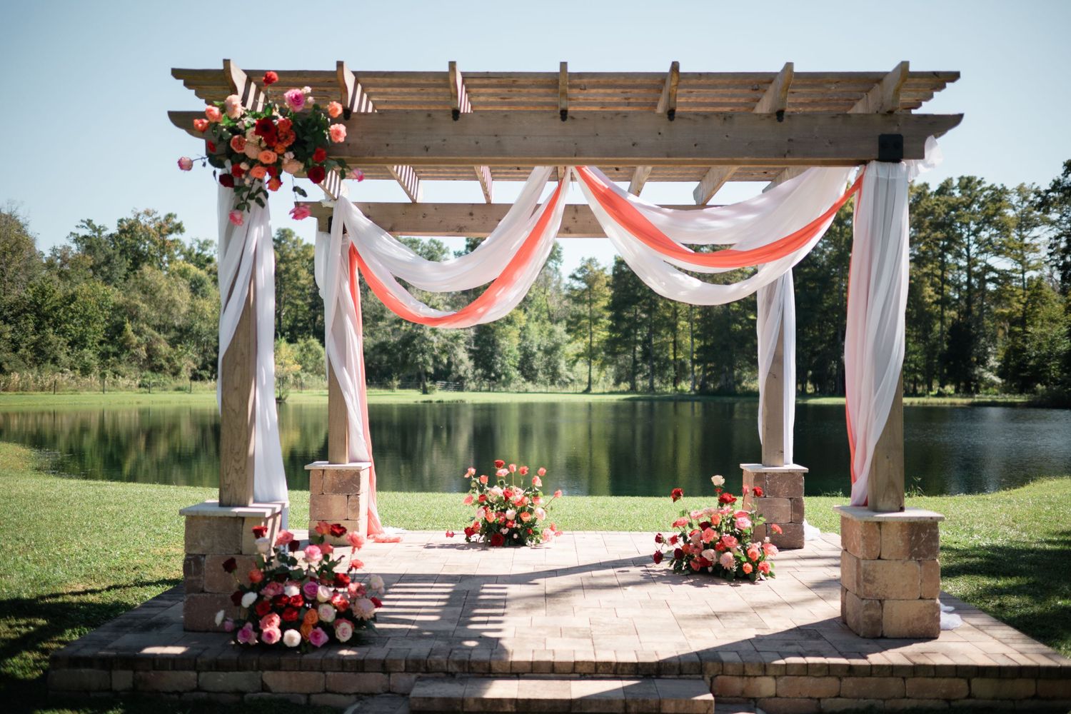 Outdoor wedding ceremony setup with white chairs and coral floral arrangements under a wooden pergola by a lake.