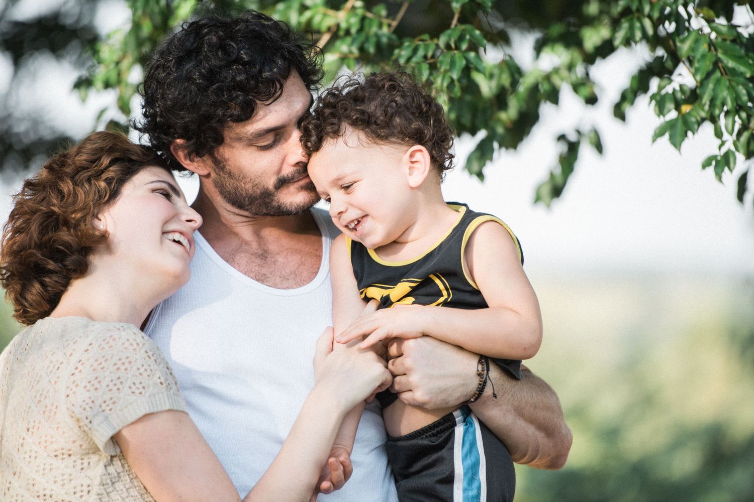 A couple shares playful moments with their baby outdoors under green trees in a series of candid photos.