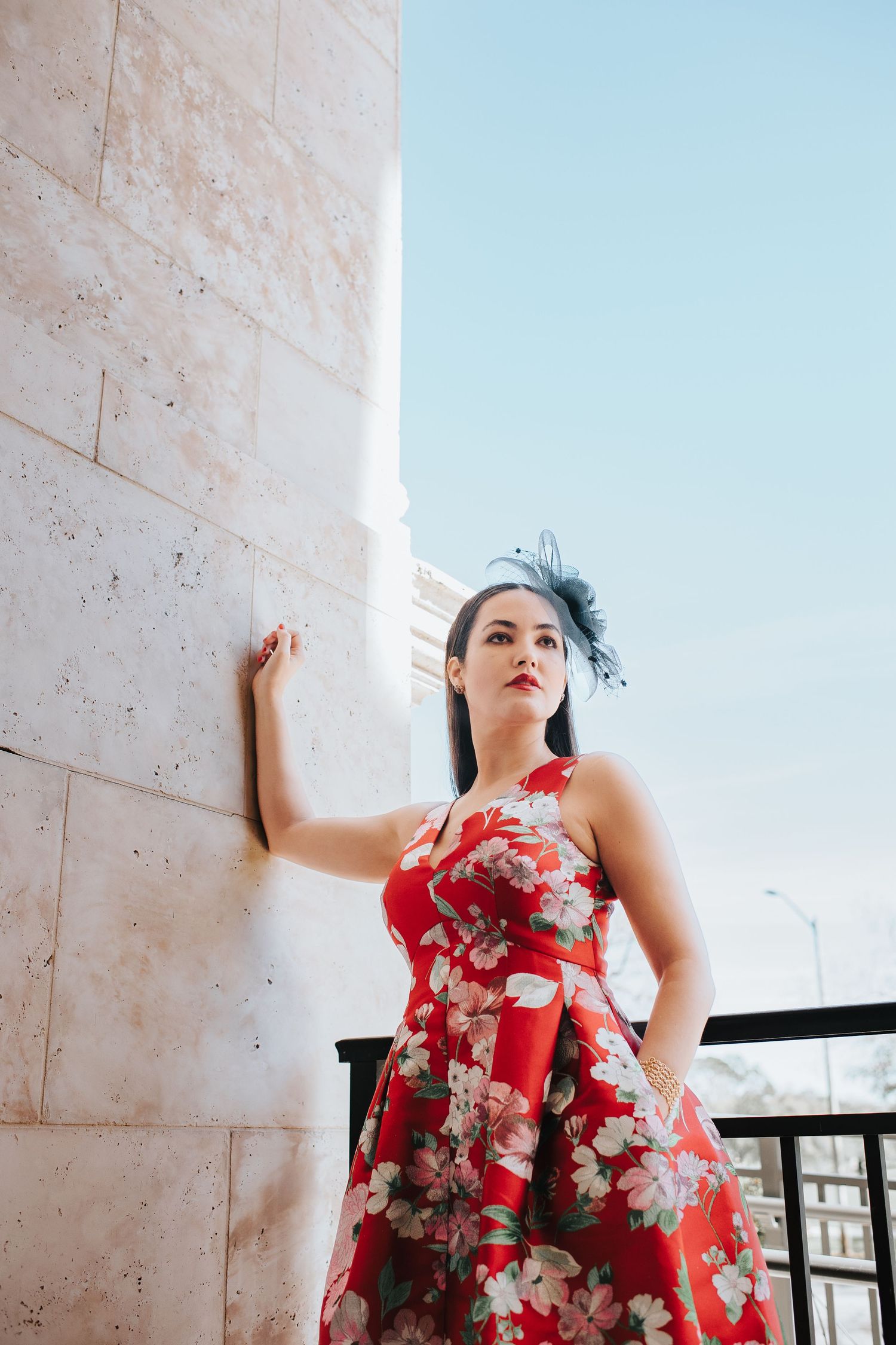 Woman in red floral dress poses against stone wall under bright blue sky.