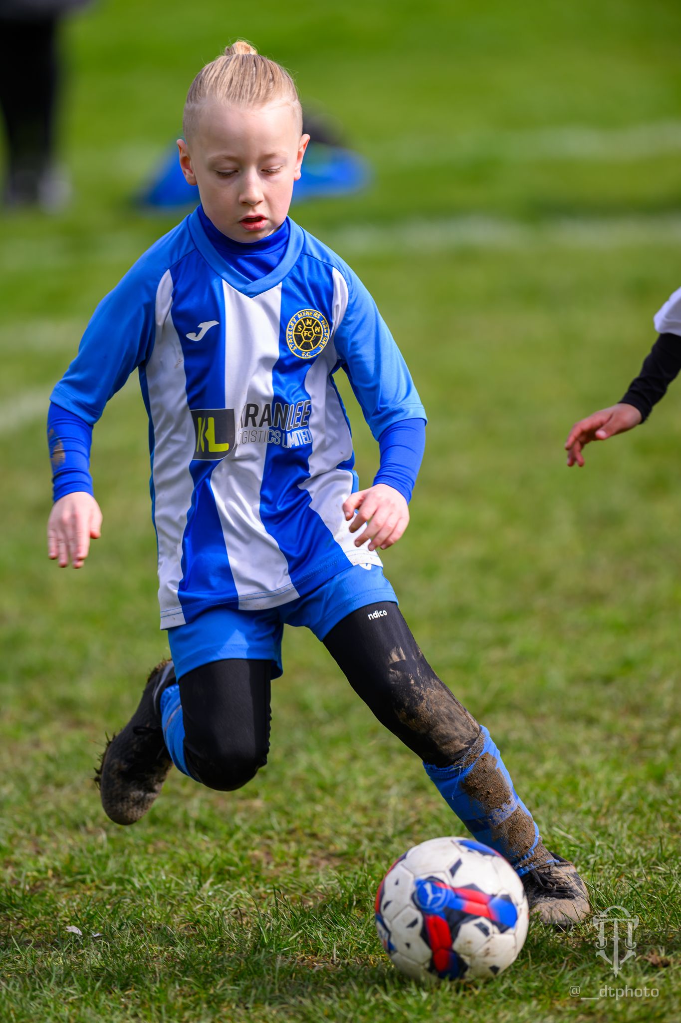 Young soccer player in blue and white striped uniform dribbling ball on muddy grass field during match.