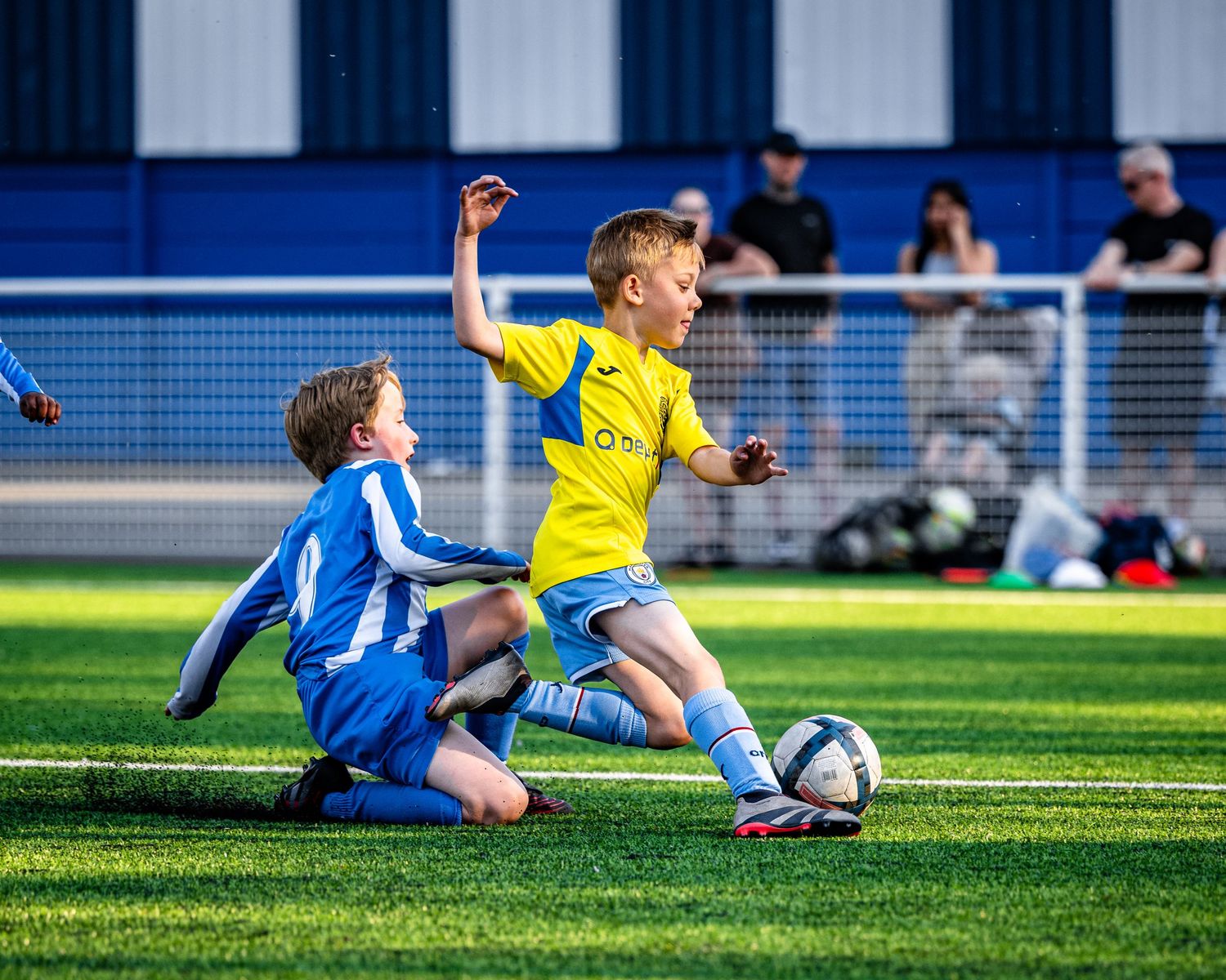 Two youth soccer players compete for the ball during an intense tackle on artificial turf field.