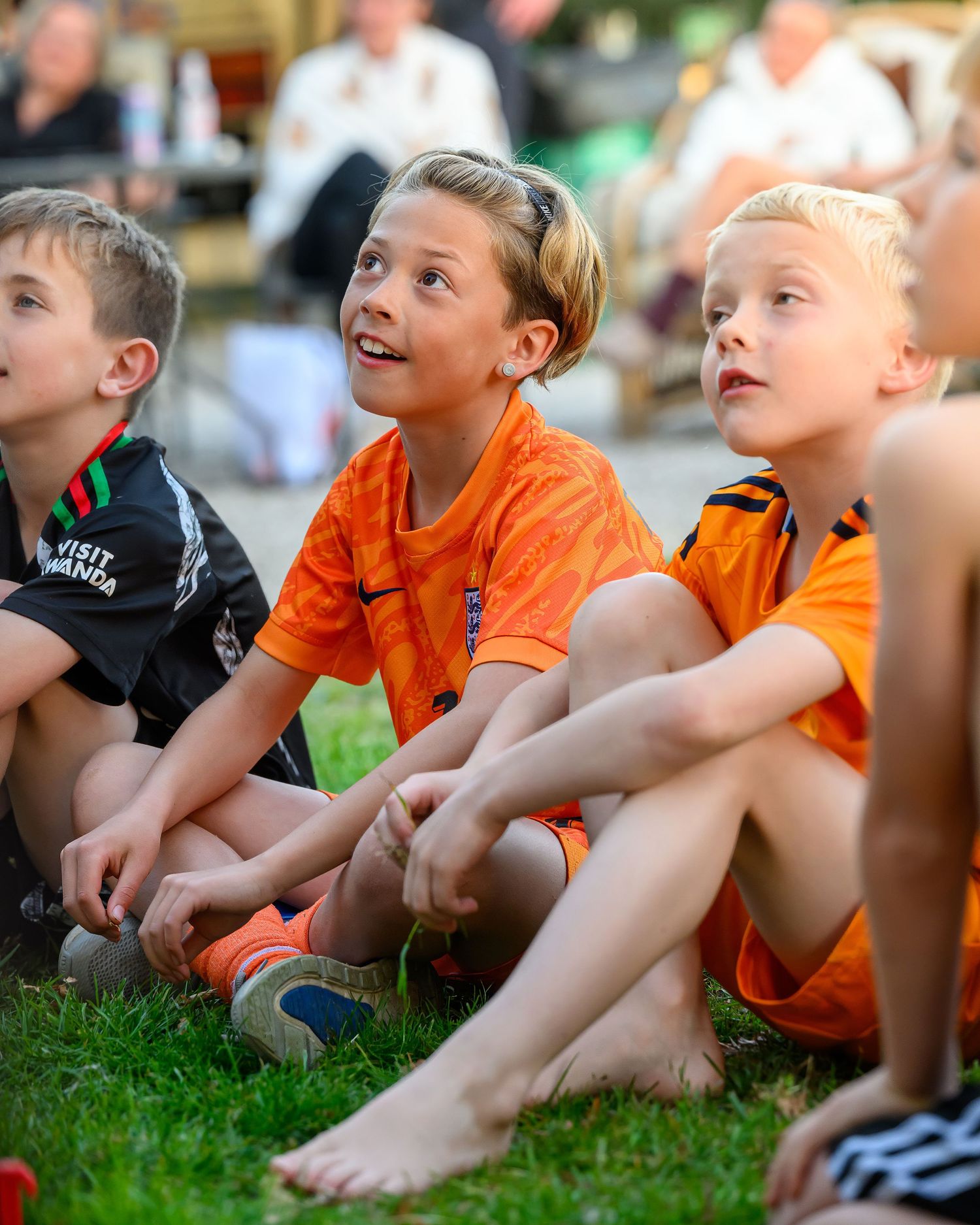 Three young spectators in orange and black clothing sit attentively on grass watching an event outdoors.
