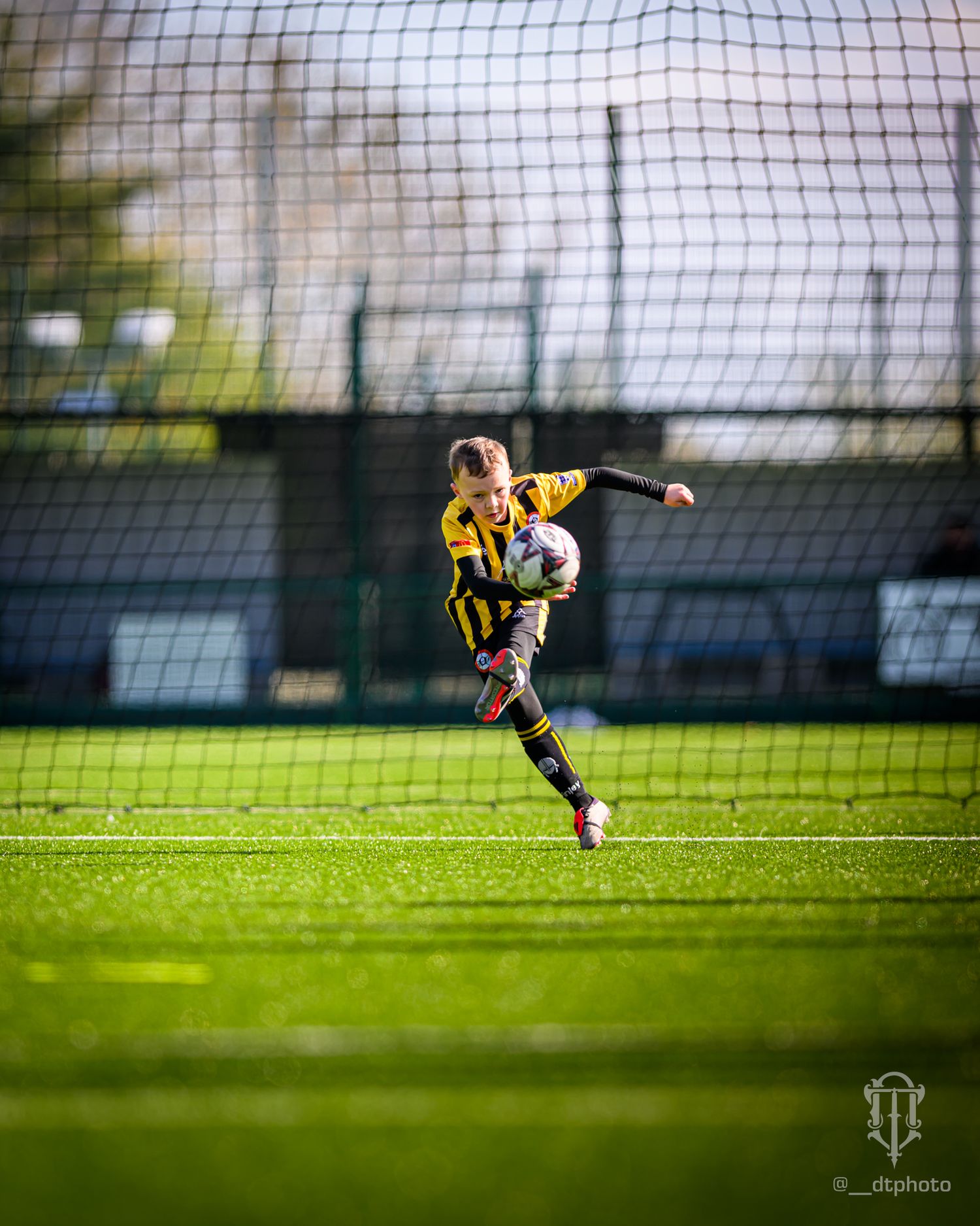 Soccer player in yellow and black striped jersey kicks the ball on green field with net visible in background.