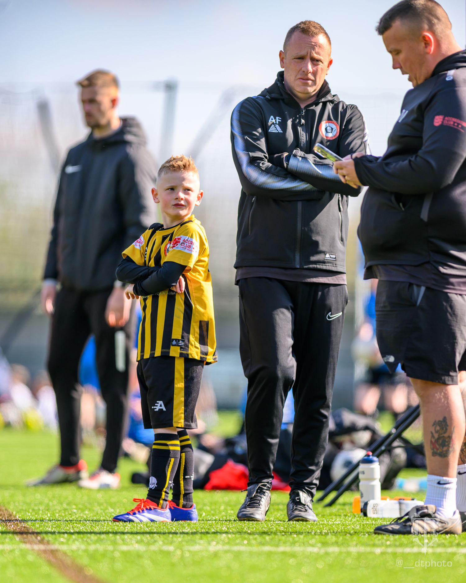Young player in yellow and black striped uniform stands with coaches during outdoor soccer training session.