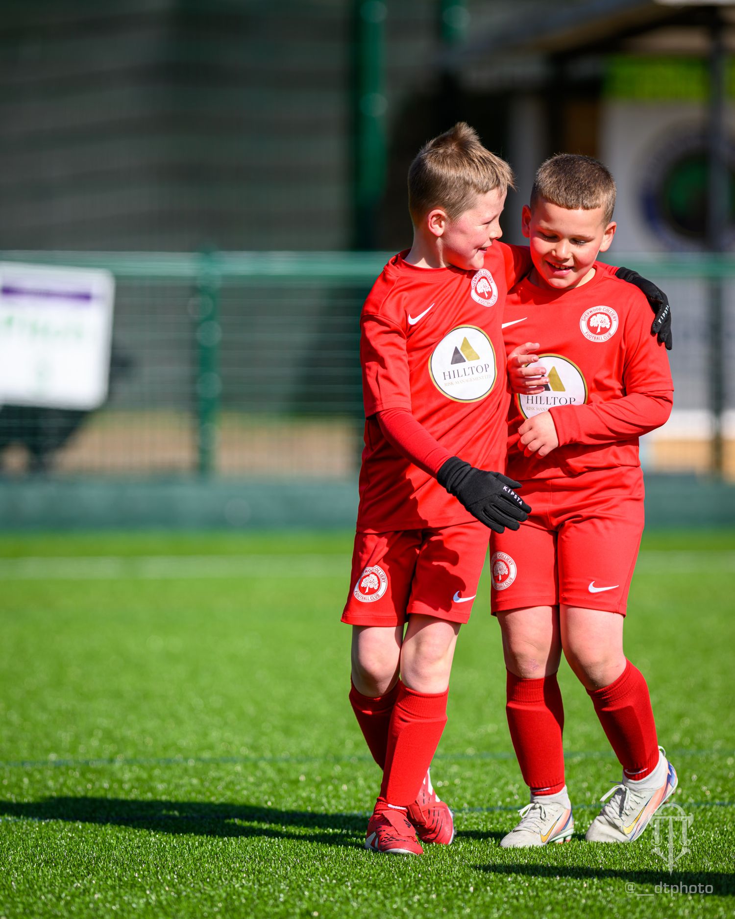 Two young soccer players in red uniforms share a lighthearted moment on a sunny grass field.