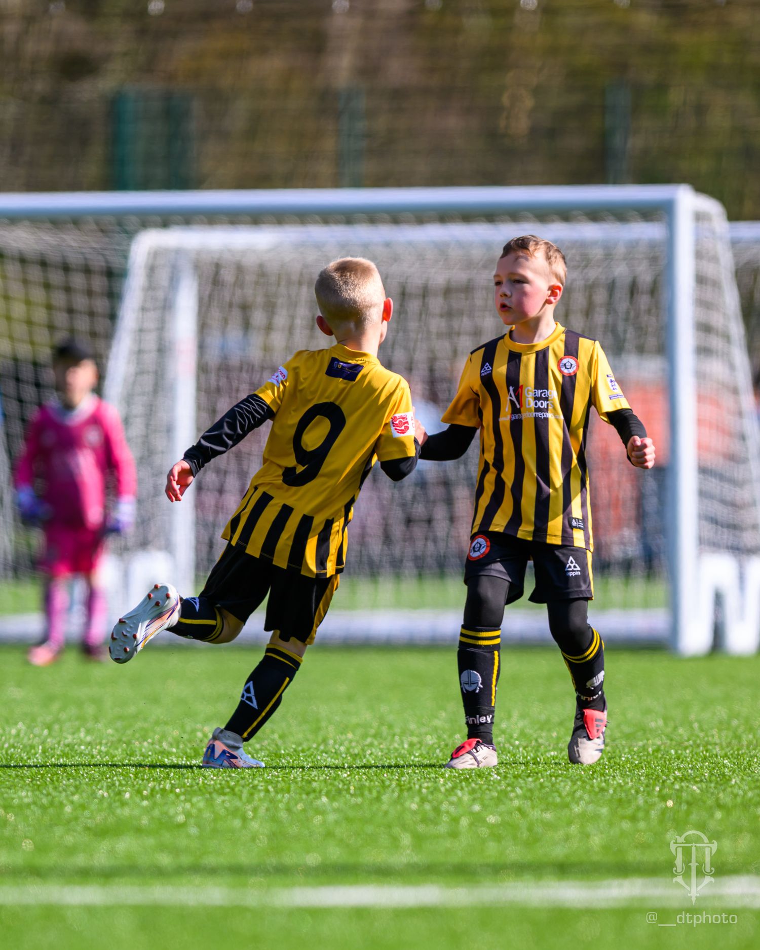 Two youth soccer players in black and yellow striped uniforms play on a grass field with a goal visible in background.