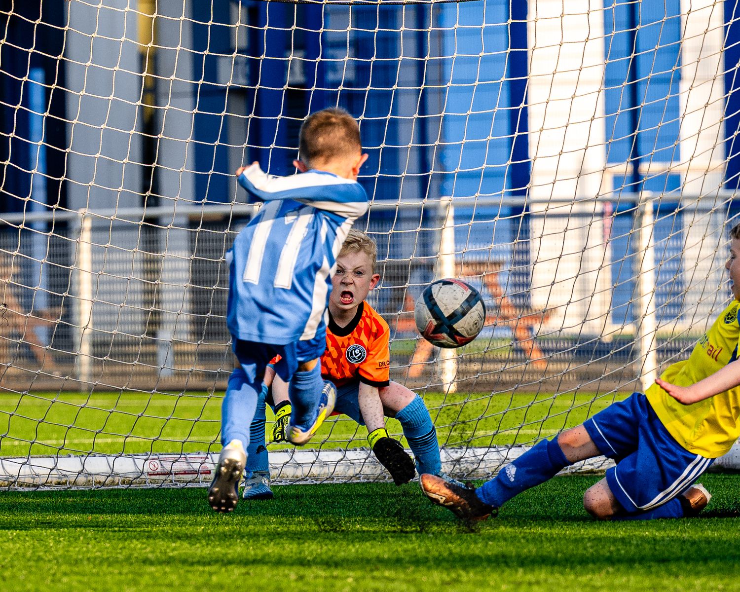 Soccer player in blue and white stripes attempts scoring against diving goalkeeper during youth match.