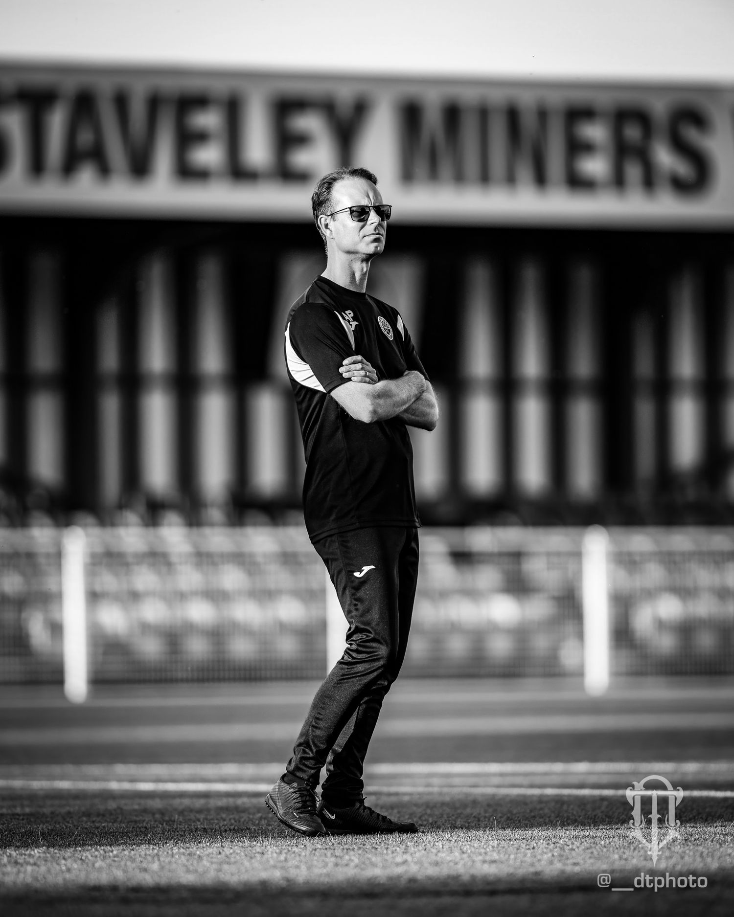 A black and white photo of a person in sunglasses and dark clothing standing on a sports field in front of stadium signage.