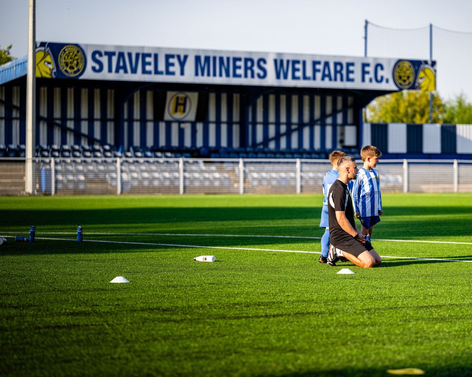 Soccer players kneel on field at Staveley Miners Welfare FC stadium with club signage visible in background.