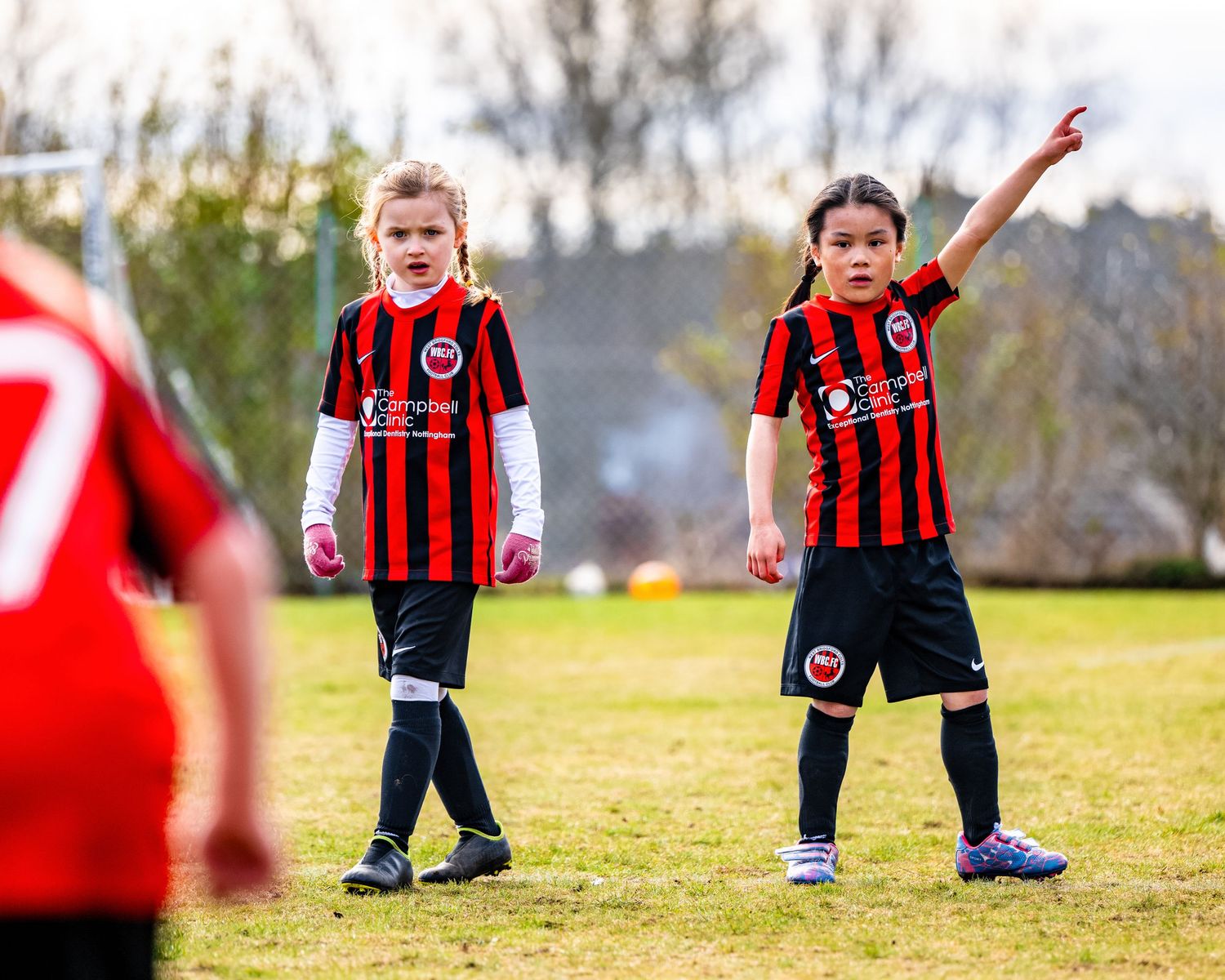 Two players in red and black striped jerseys gesture during a youth football match on a sunny field.