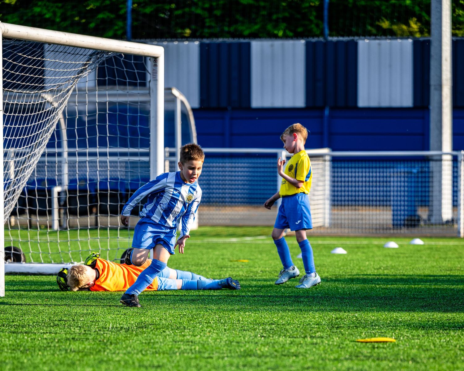 Soccer player in blue and white striped jersey scores goal during competitive match on sunny afternoon.