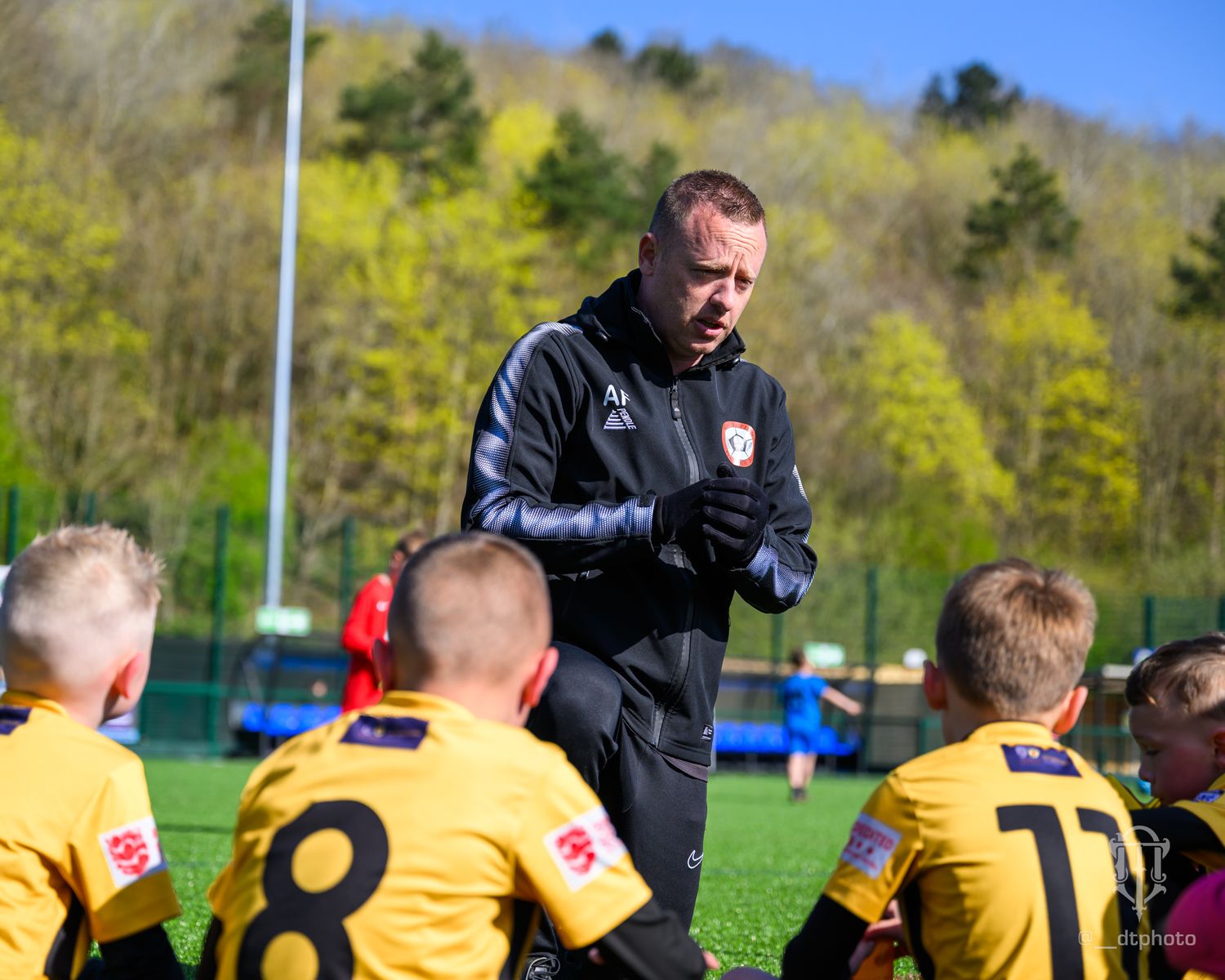 Youth sports coach in black jacket gives instructions to players in yellow jerseys during outdoor practice session.