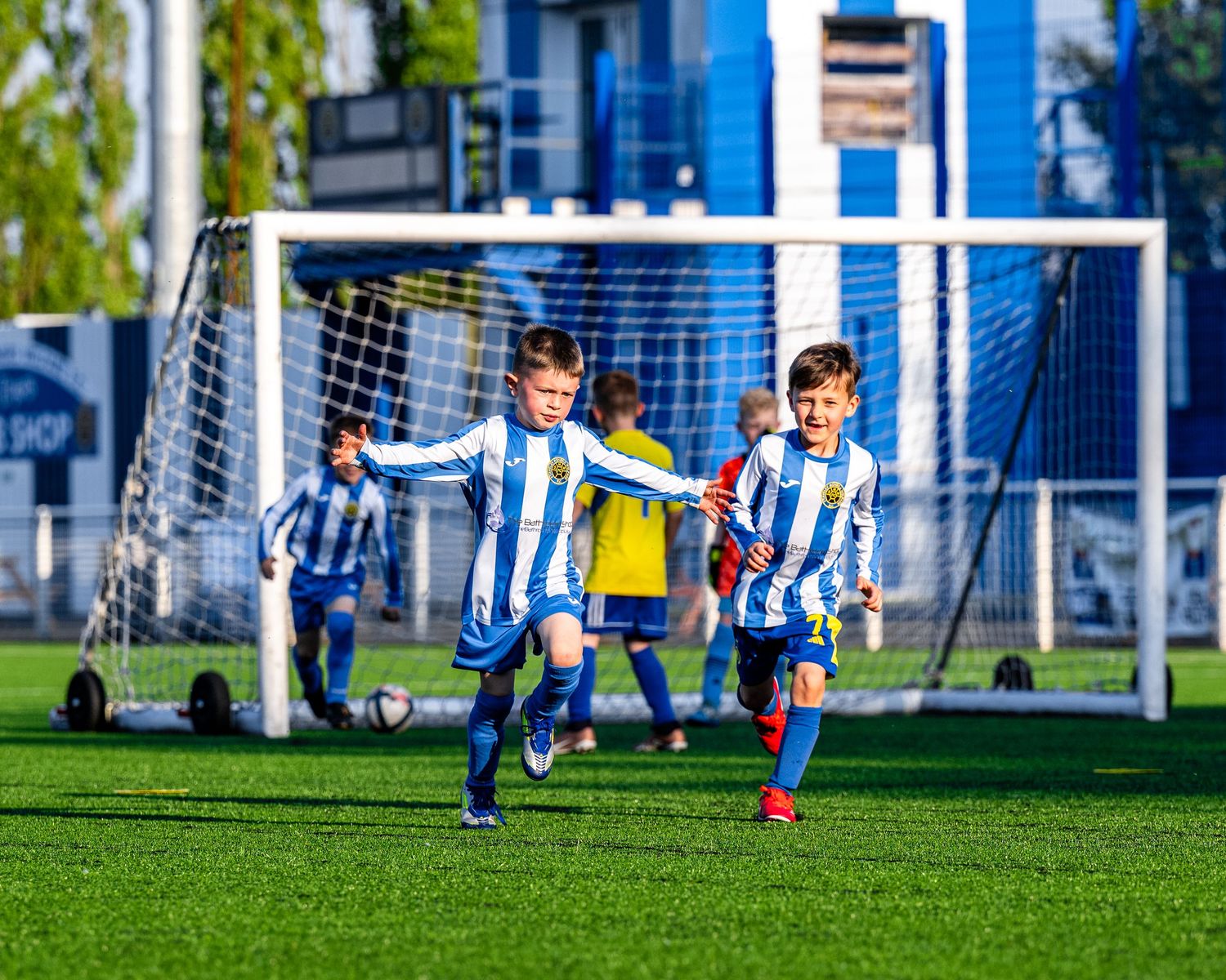Young soccer players in blue and white striped jerseys playing on a sunny field with a goal net in the background.