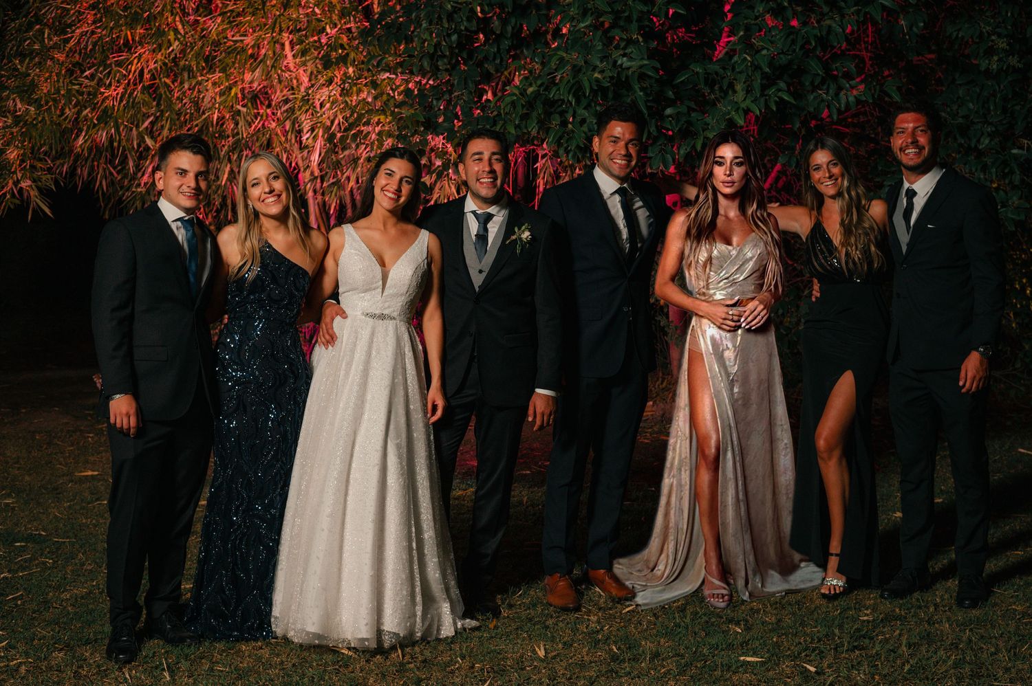 A wedding party gathers at dusk for group photos in a natural outdoor setting.